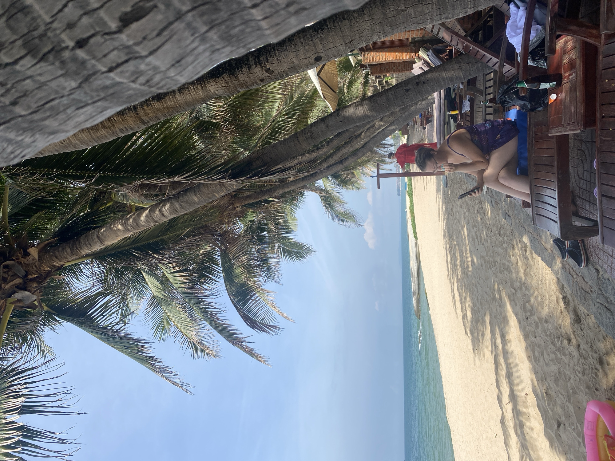 A woman is sitting on a lounge chair on a sandy beach, looking at her phone. She is wearing a purple swimsuit. Behind her, there are palm trees and a building. The ocean is in the background. The scene appears to be in Phu Thuy Ward, Vietnam.