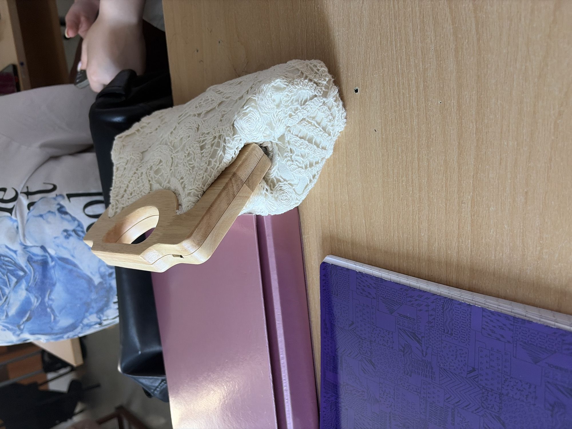 The image shows a light wooden desk with a light-colored wooden surface. A white, crocheted purse with a wooden handle is placed on the desk. Underneath the purse, two notebooks are stacked. The top notebook is a light pink color, and the bottom notebook is a deep purple color with a geometric design. The image is taken from an overhead perspective. In the background, a person is sitting on a chair.
There is no information to indicate that this image was taken in Burgas, Bulgaria.