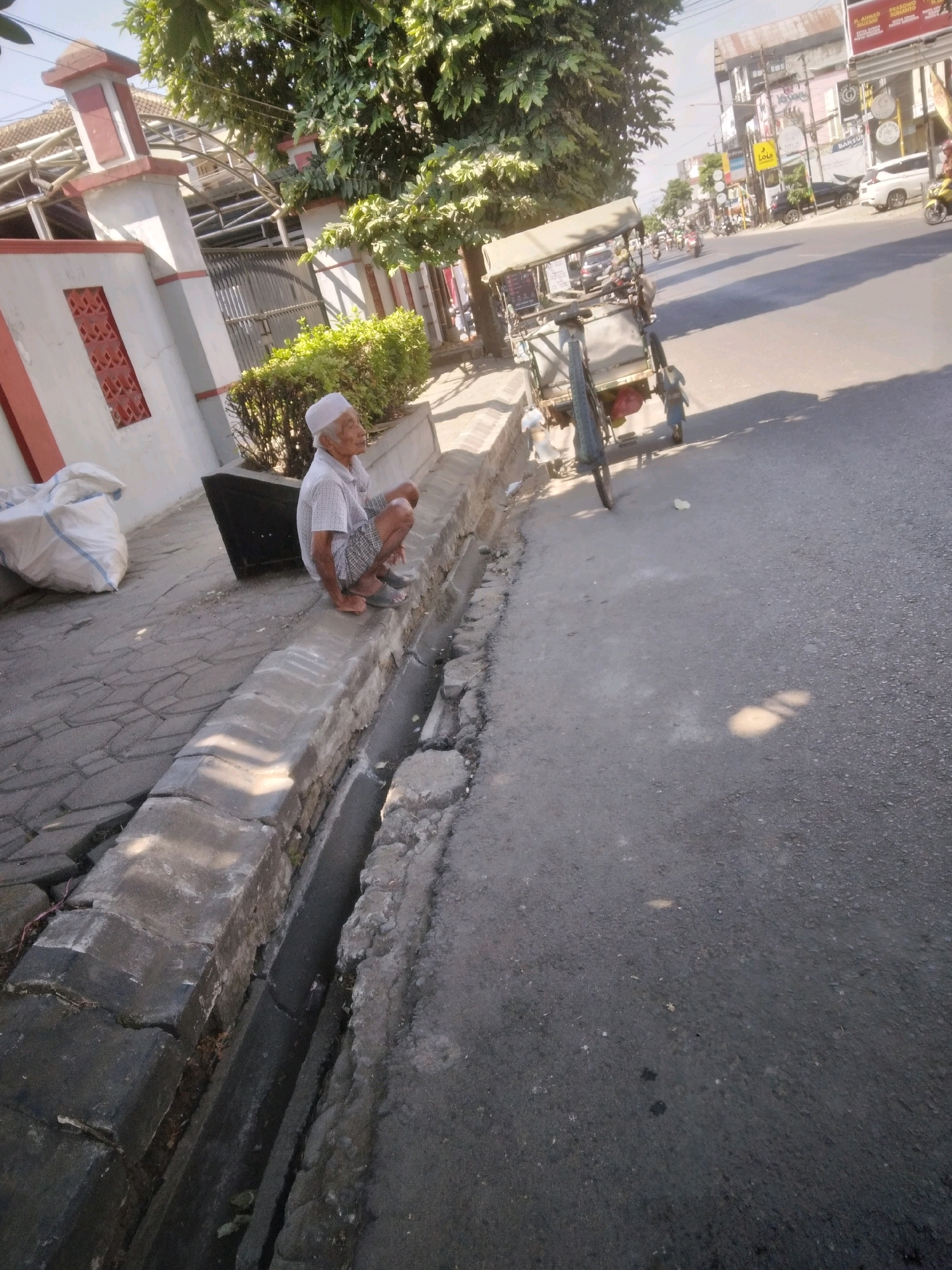 An elderly man wearing a white cap and a plaid shirt is crouching on a concrete gutter next to a paved road in Sumampir, Indonesia. The man is facing to the right, with his left hand resting on his knee and his right hand near his right foot. A white plastic bag is visible behind him on the paved sidewalk. A rickshaw is parked on the left side of the road, facing left. The road is lined with trees and buildings, and other vehicles and pedestrians are visible in the background, suggesting a busy urban environment. The sky is clear and the sun is shining.