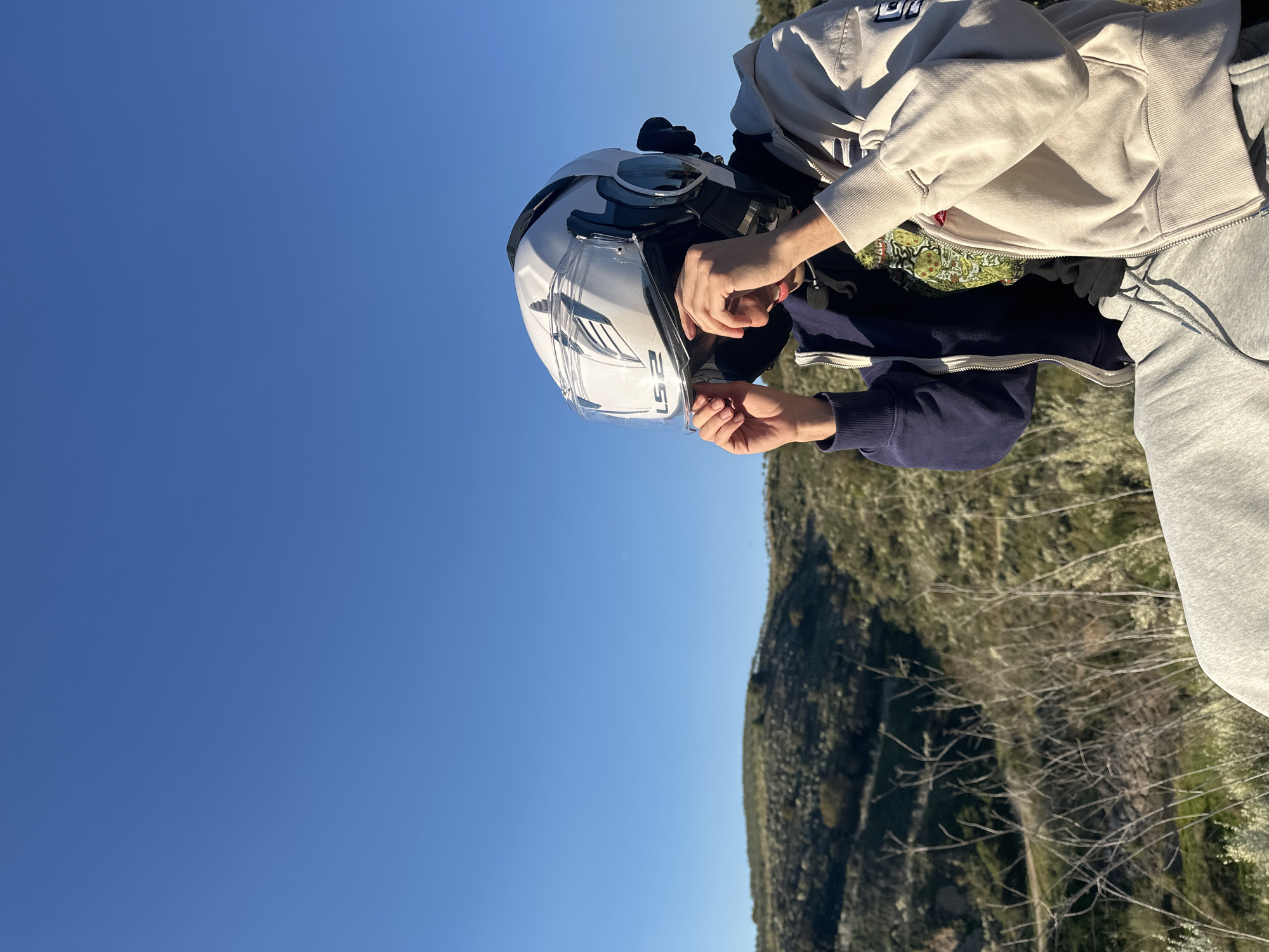 A person wearing a white motorcycle helmet is depicted with hands adjusting the visor. The individual is wearing a combination of outerwear: a light-colored zip-up hoodie, a navy blue zip-up jacket, and light gray sweatpants. The backdrop is a clear blue sky, with a tree covered hill behind. The location is identified as Mirandela, Portugal.