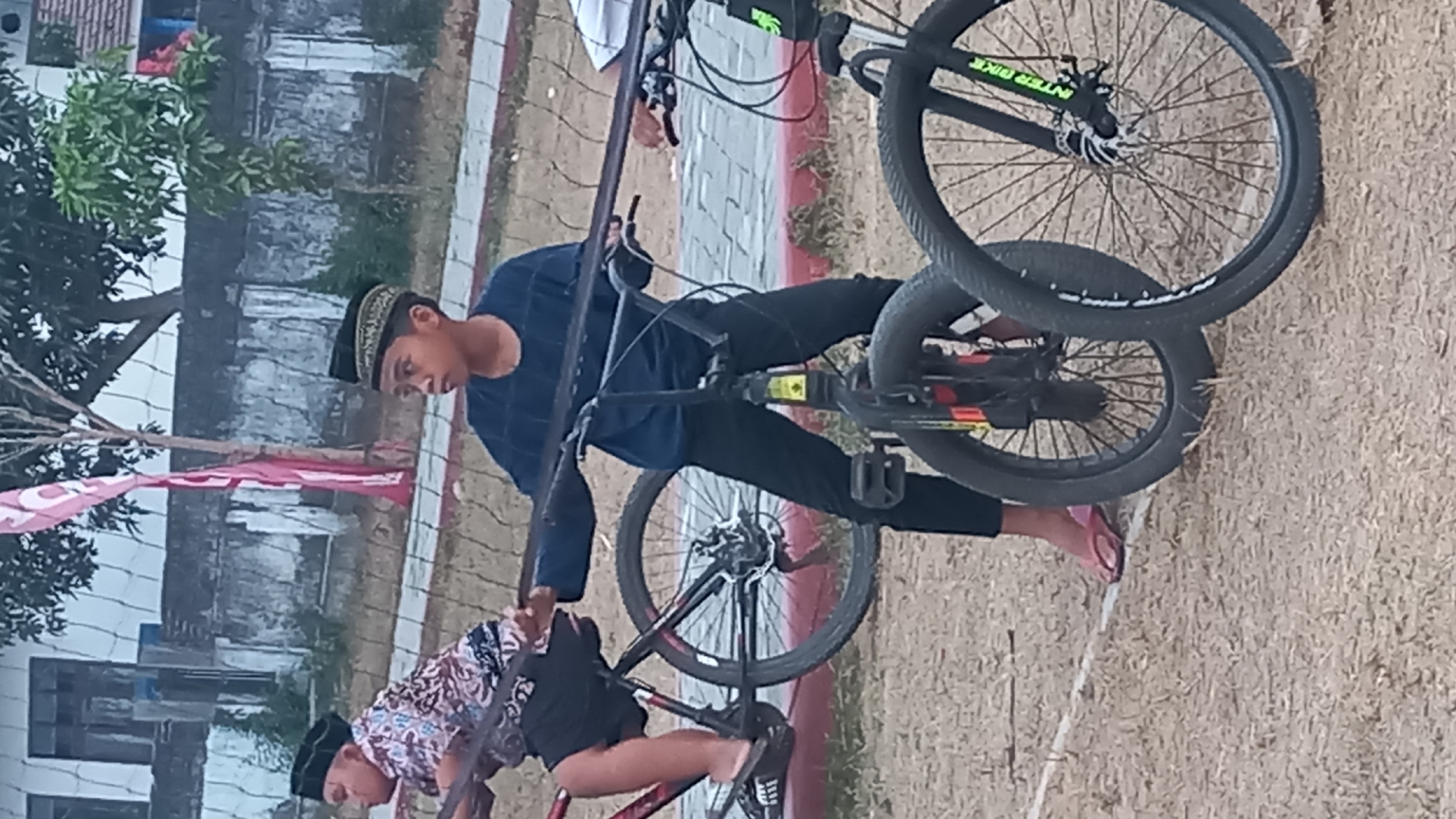 Two young boys, one on a red bicycle and another on a black BMX-style bicycle, are standing next to each other. Both boys are wearing traditional Indonesian black hats, called peci. The boy on the left is wearing a patterned shirt and dark shorts. The boy on the right is wearing a dark blue long-sleeved shirt and black pants. The BMX bicycle has a yellow sticker on the frame. They are standing on a dirt ground with dry grass and a white line marking a boundary. In the background, there is a low brick wall and some trees. A pink flag is visible on a pole in the background. The scene appears to be an outdoor recreational area. This location is likely within the city of Pare, Indonesia, given the cultural context implied by the attire and the general setting.
