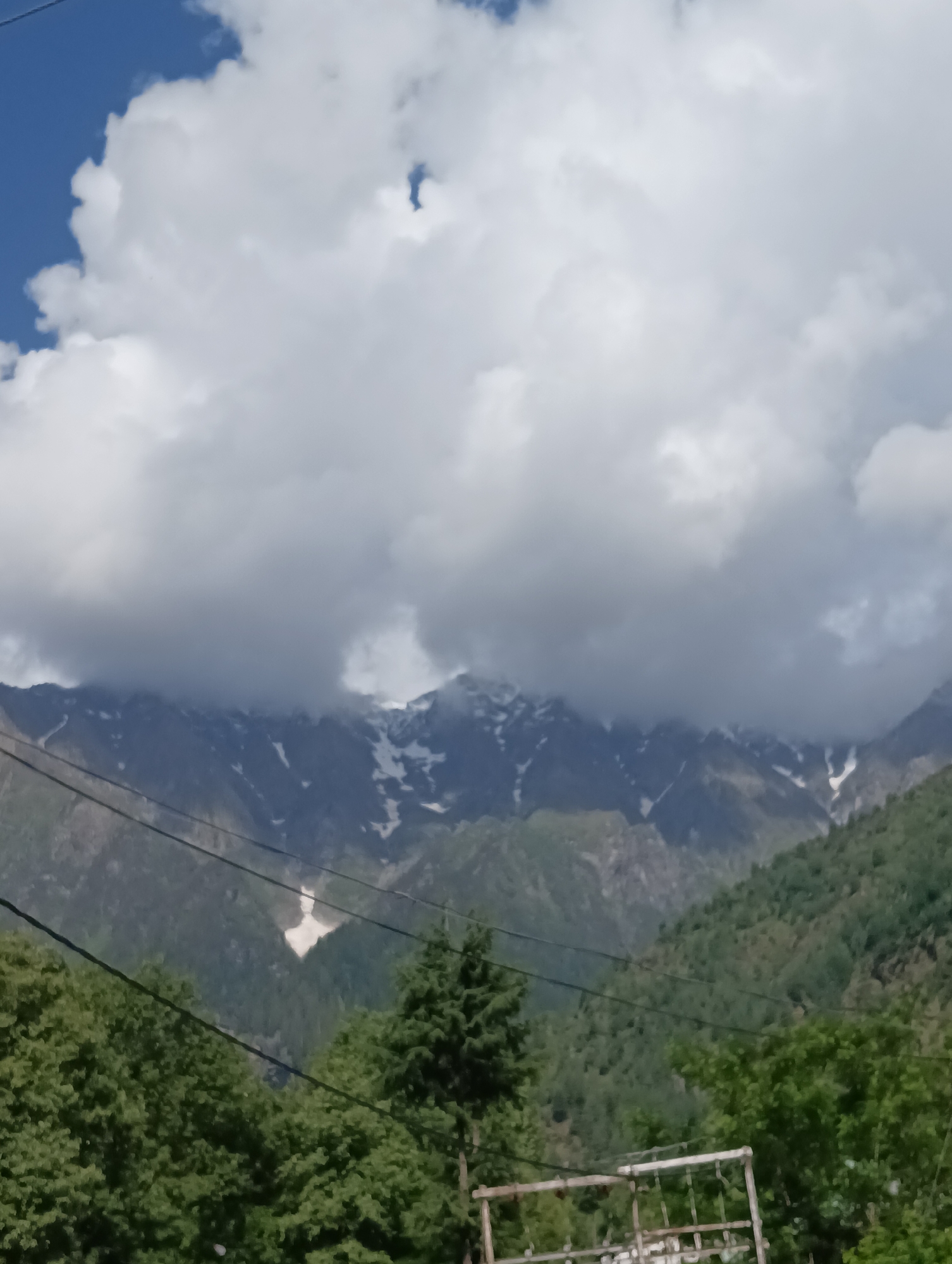 The image shows a mountain range partially obscured by clouds. The lower slopes of the mountains are covered in dense green vegetation, with some patches of snow visible higher up. In the foreground, there are trees and power lines. The sky above is a mixture of blue and white clouds. The scene suggests a natural, remote environment, possibly in a mountainous region. Without further information or context, pinpointing the exact location within Barheta, India, is not possible. The image is a still photograph capturing a natural landscape.