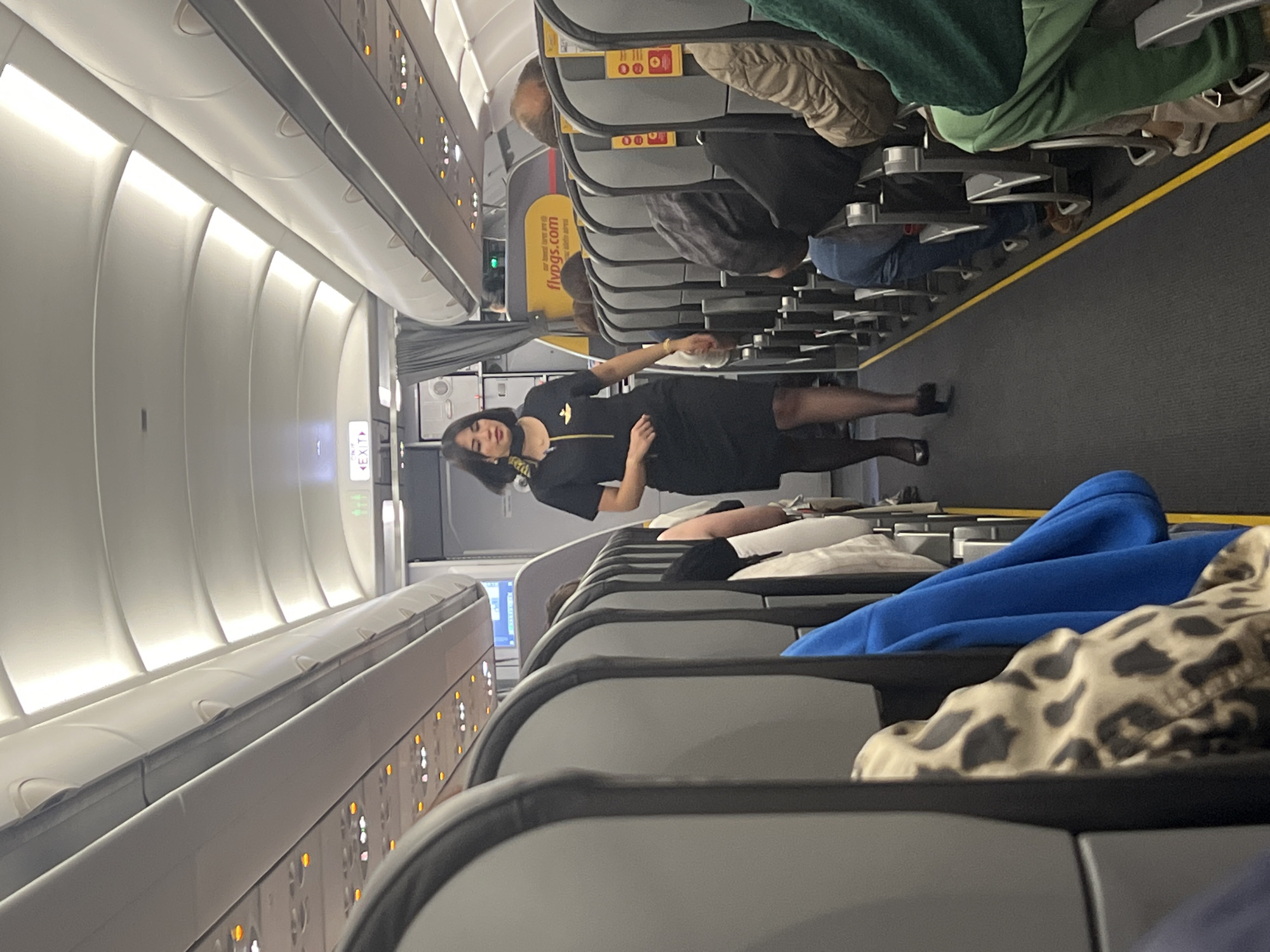 The image displays the interior of a commercial airplane cabin, specifically an economy class section. The main subject is a female flight attendant, dressed in a black uniform with yellow trim and a golden emblem, standing in the aisle. She has dark hair and is looking towards the camera.
The setting shows rows of grey passenger seats with yellow accents lining both sides of the aisle. Several passengers are visible, mostly from the back or side, seated in their seats. Some seats feature items like a green blanket, a blue hooded garment, and a leopard-print blanket, suggesting passengers are settled for the flight. Overhead bins are closed above the seats, and passenger service units with lights and call buttons are visible. A yellow safety stripe runs along the floor of the aisle.
The primary activity depicted is a flight attendant performing duties or pausing in the aisle while passengers are seated. The overall atmosphere appears calm and orderly, typical of a flight either during boarding, cruising, or deplaning.
Notable details include the airline branding "flypgs.com" visible on a yellow panel, identifying the carrier as Pegasus Airlines. An "EXIT" sign with an arrow is also visible on the cabin wall. The cabin is brightly lit by artificial lighting. Given the provided context of Saint Petersburg, Russia, this is likely a Pegasus Airlines flight to or from that location.