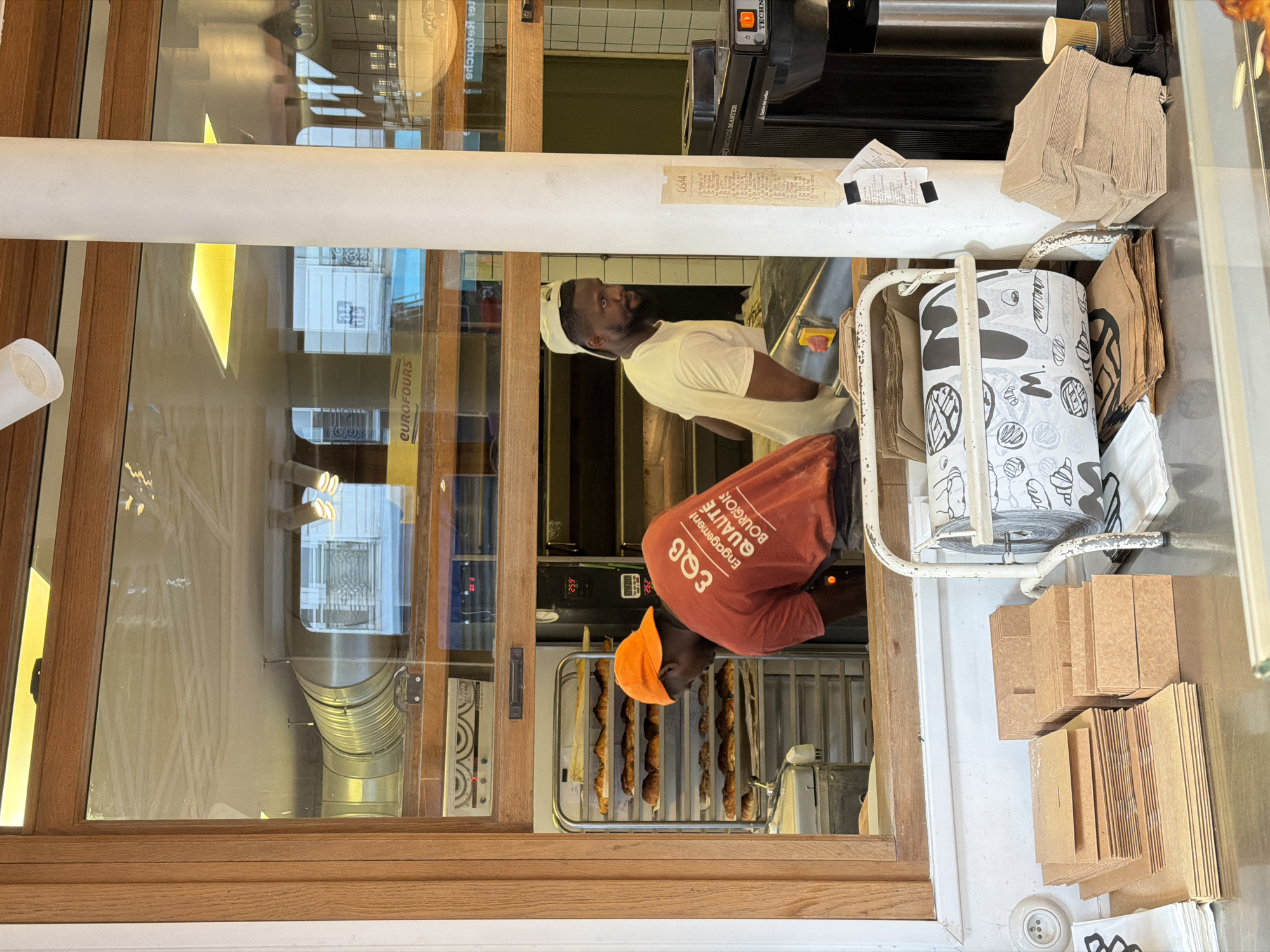 Here's a detailed description of the image:
**Scene:** The image appears to be taken from outside a bakery or pastry shop in Paris, France, looking in through a large window. The window is divided into panes with wooden frames.
**People:** There are two Black men visible inside the shop:
* One man is wearing an orange cap and a red-orange t-shirt with white text that reads "EQB Engagement QUALITÉ BOURGIOIS”. He appears to be taking baked goods from an oven.
* The second man is wearing a white cap and a white shirt and appears to be assisting with the baking or sales process.
**Objects:** The scene contains various objects related to a bakery:
* An oven with several racks of baguettes or similar bread.
* A cart with brown paper bags and wrappers with bakery-themed designs.
* Stacks of folded paper bags.
* A black TECHN IMOD CAMASTER oven.
* A white support beam with handwritten text on it.
* Reflection of buildings in window.
* The window sticker EUROFOURS.
**Actions and Interactions:**
* The man in the orange cap appears to be removing bread from the oven.
* The other man seems to be involved in food preparation or packaging, likely interacting with the first man.
**Environment:**
* The interior has a commercial bakery aesthetic.
* The exterior is likely a street in Paris.
**Additional details:**
* The text on the white beam reads "0644"
* The oven has digital timer indicators that read '.652', '14.00' and '292'.