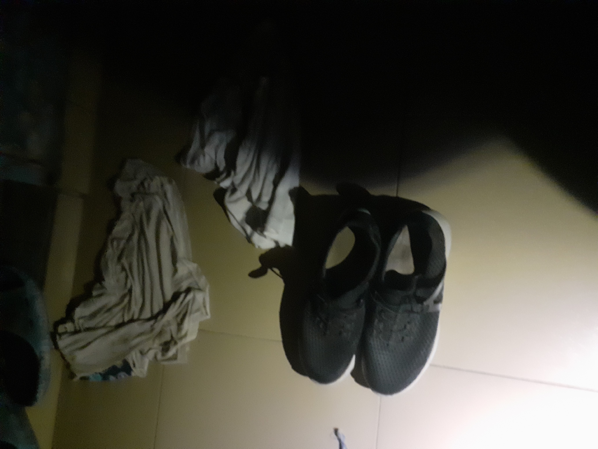 A pair of black athletic shoes are placed on a light-colored tiled floor. Above the shoes, there are piles of folded clothing, appearing to be light-colored fabric. The scene is dimly lit, with shadows obscuring parts of the environment. There is no indication of the location being in Jalingo, Nigeria.