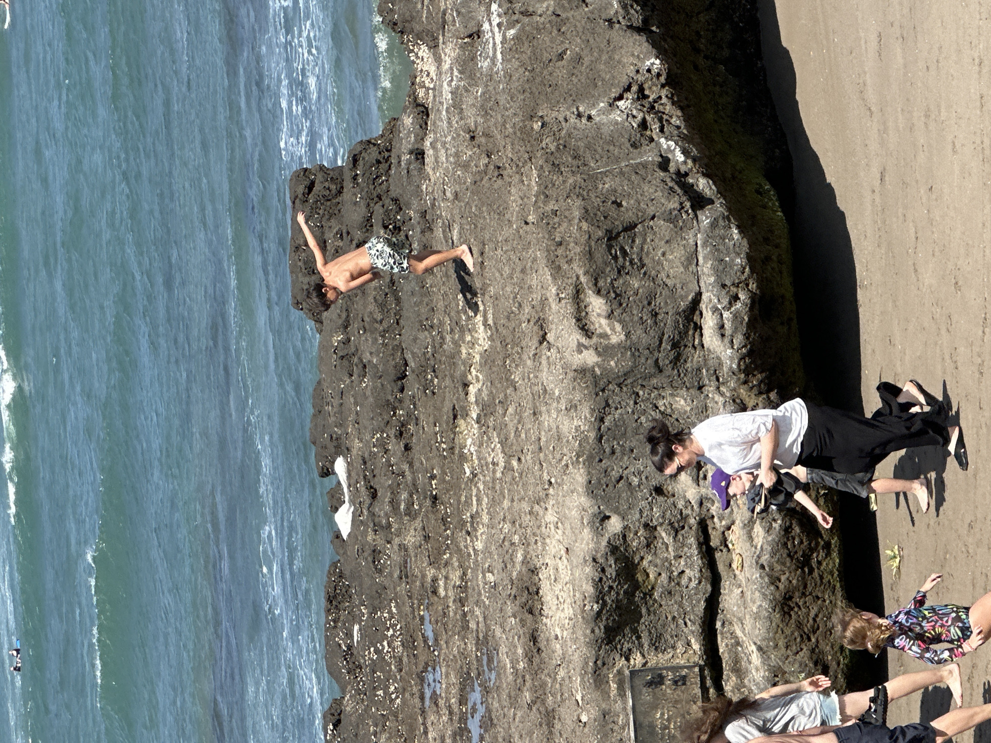The image shows a group of people on a rocky coastline next to the ocean. The water is a deep blue and appears to be quite wavy. The rocks are dark and rugged, with some patches of green moss.
In the foreground, a person in a white shirt and black pants is lying on their side on the sand, facing the ocean. Next to them, another person in a white shirt and black shorts is also lying down. Further down the beach, a young person in a colorful swimsuit is standing. In the background, on the rocks, a young person is lying on their stomach, looking out at the water.
The sun appears to be high in the sky, suggesting it is daytime. The weather seems to be warm and sunny, as people are dressed in summer clothing. The scene appears to be a relaxed day at the beach, with people sunbathing and enjoying the ocean views.
The location is identified as North Kuta, Indonesia. There is no visible text in the image.