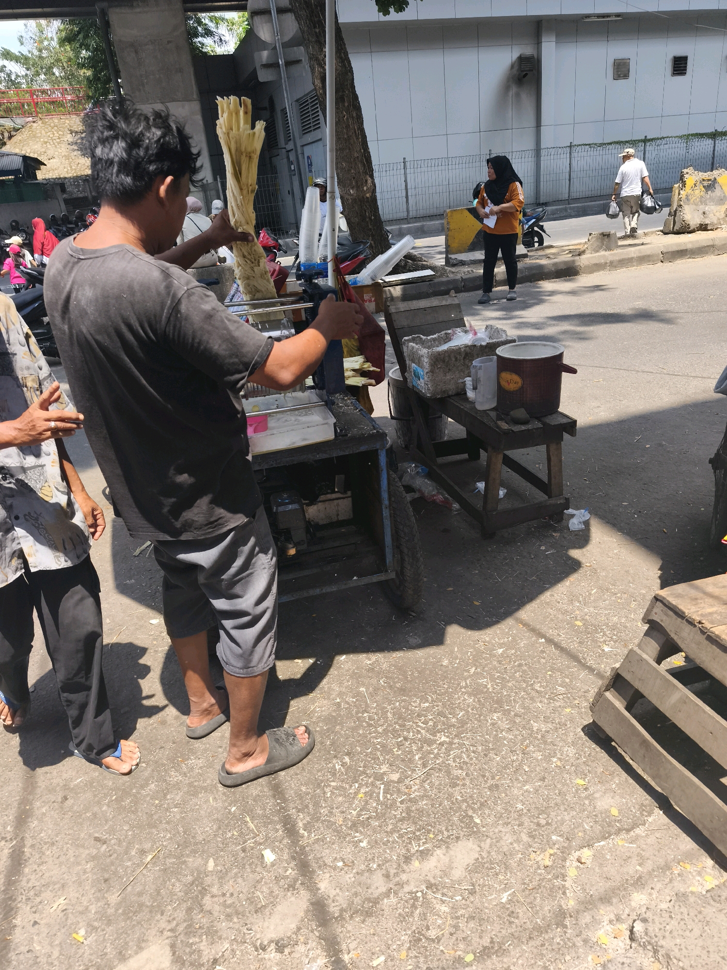 In Kelurahan 16 Ilir, Kec. Ilir Timur I, Indonesia, a man with dark, messy hair is operating a sugarcane juice machine on a street. He is wearing a dark grey t-shirt and grey shorts, and dark sandals. He is holding a bundle of sugarcane stalks, preparing to feed them into the machine. To his left, another person, partially visible, is wearing a patterned short-sleeved shirt and black pants. The sugarcane juicing machine is mounted on a cart with wheels and appears to be powered by a small engine visible underneath. Nearby, a wooden stand holds a large red cooler, a pitcher, and a rectangular ice chest filled with ice. In the background, two women are walking on the sidewalk; one is wearing a black hijab and an orange shirt, holding a piece of paper, while the other is an elderly person in a white shirt and hat. Various vehicles, including motorcycles, are parked along the street. The scene is illuminated by bright sunlight, casting strong shadows on the asphalt road. The environment appears to be an urban street with buildings and overhead structures visible.