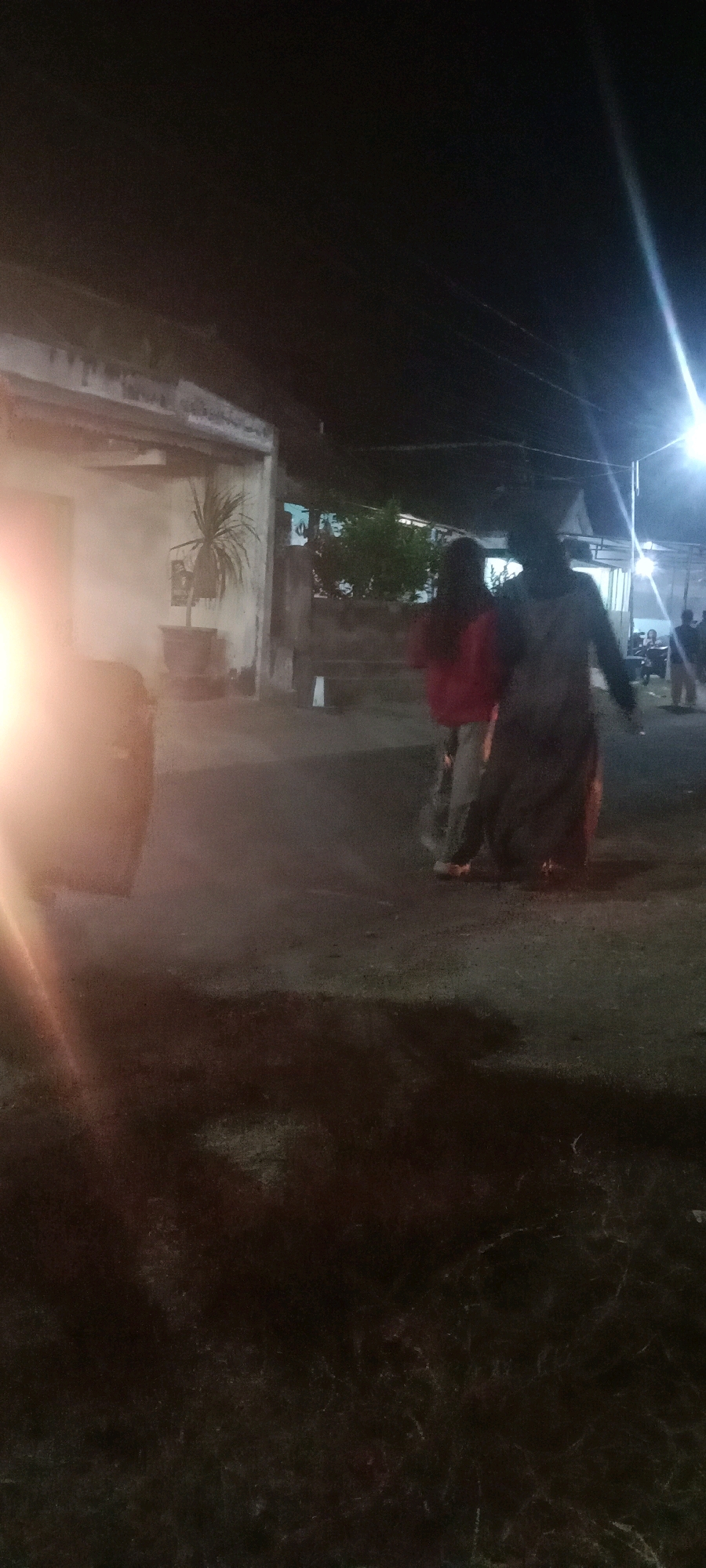 The image depicts a nocturnal scene in what appears to be a residential area in Kras, Indonesia, potentially a street or courtyard. The lighting is dim, with a bright light source from the right, possibly a street lamp, casting a strong glare and illuminating the area. In the foreground, on the left, a large, opaque object, likely a drum or a trash bin, is partially visible and heavily lit.
In the middle ground, two individuals are walking away from the camera. The person on the left is wearing a red top and appears to be holding onto the person on the right. The individual on the right is wearing a dark, loose-fitting garment, possibly a long dress or robe, and is also walking away. Their interaction is not clearly discernible due to the low light and distance. Behind them, a building with a textured, light-colored wall is visible, with a potted plant featuring long, slender leaves placed near its entrance. Further back, more buildings and vegetation are faintly visible, suggesting a built-up environment. The ground is a dark, uneven surface, possibly asphalt or packed earth, with some patches of dry grass or weeds in the lower part of the frame. The overall atmosphere is dark and somewhat hazy due to the lighting conditions.