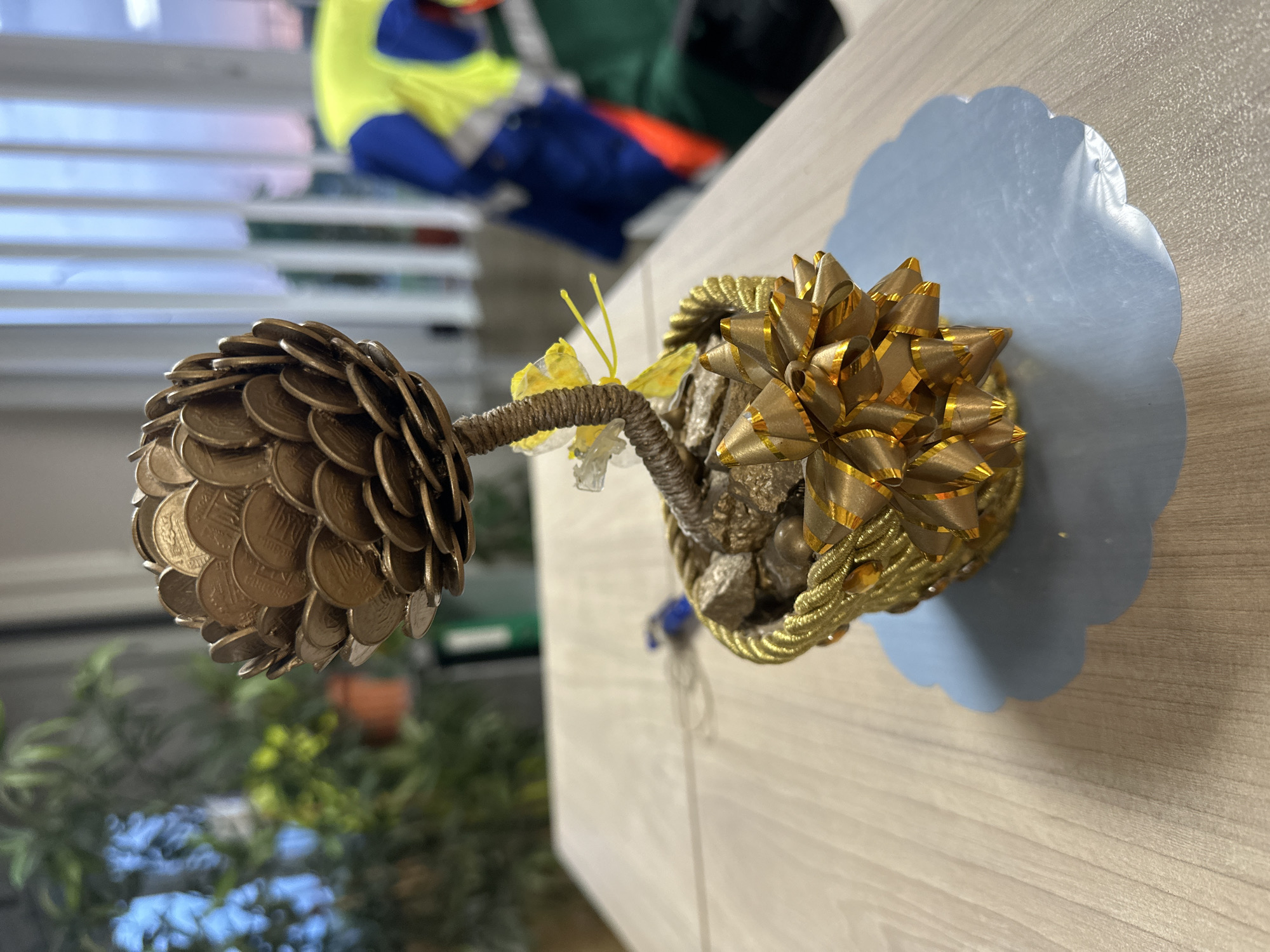 The image shows a close-up, top-down view of a decorative object resembling a pinecone, crafted from stacked coins, likely as a monetary gift or a good luck charm. The object is positioned on a light-colored wooden surface, possibly a desk or table. The "pinecone" has a metallic sheen, appearing to be made of bronze or copper-colored coins. It is attached to a stem, which is wrapped in twine and adorned with small, rough stones and a golden ribbon bow. A delicate yellow butterfly or moth decoration with antennae is attached to the stem. The object rests on a pale blue, scalloped decorative mat. In the blurred background, a person wearing a bright yellow vest and blue clothing is visible, suggesting an indoor setting, possibly an office or workshop. The overall impression is one of a handmade, symbolic gift.