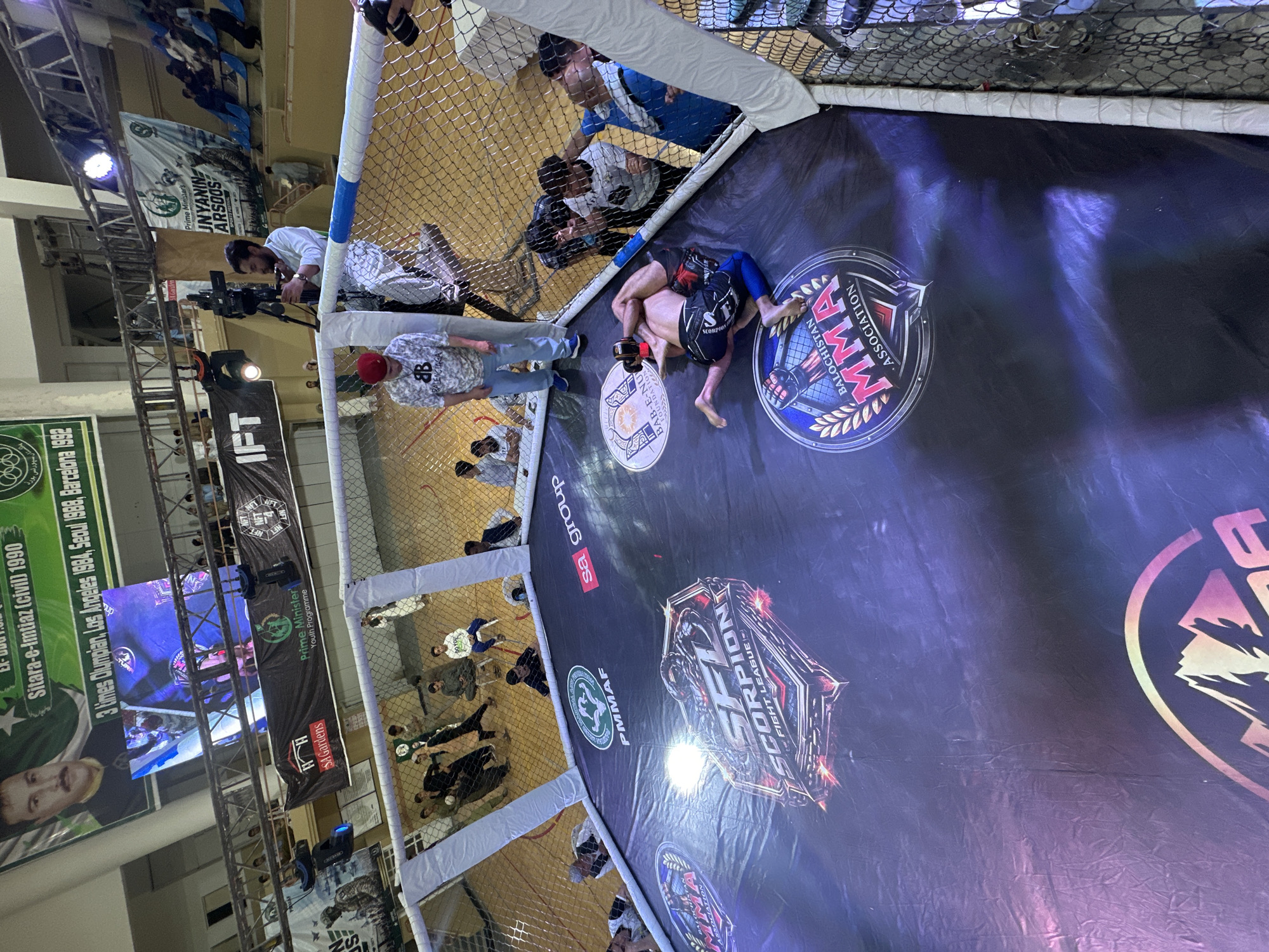 The image shows a mixed martial arts (MMA) fight taking place inside a caged octagon, likely in Quetta City Tehsil, Pakistan. Two fighters are grappling on the mat, surrounded by spectators and personnel. Banners and logos around the arena promote various organizations and sponsors, including "Balochistan MMA Association," "Scorpion Fight League," and "Prime Minister's Youth Programme." There is a cameraman filming, and a group of people are watching.