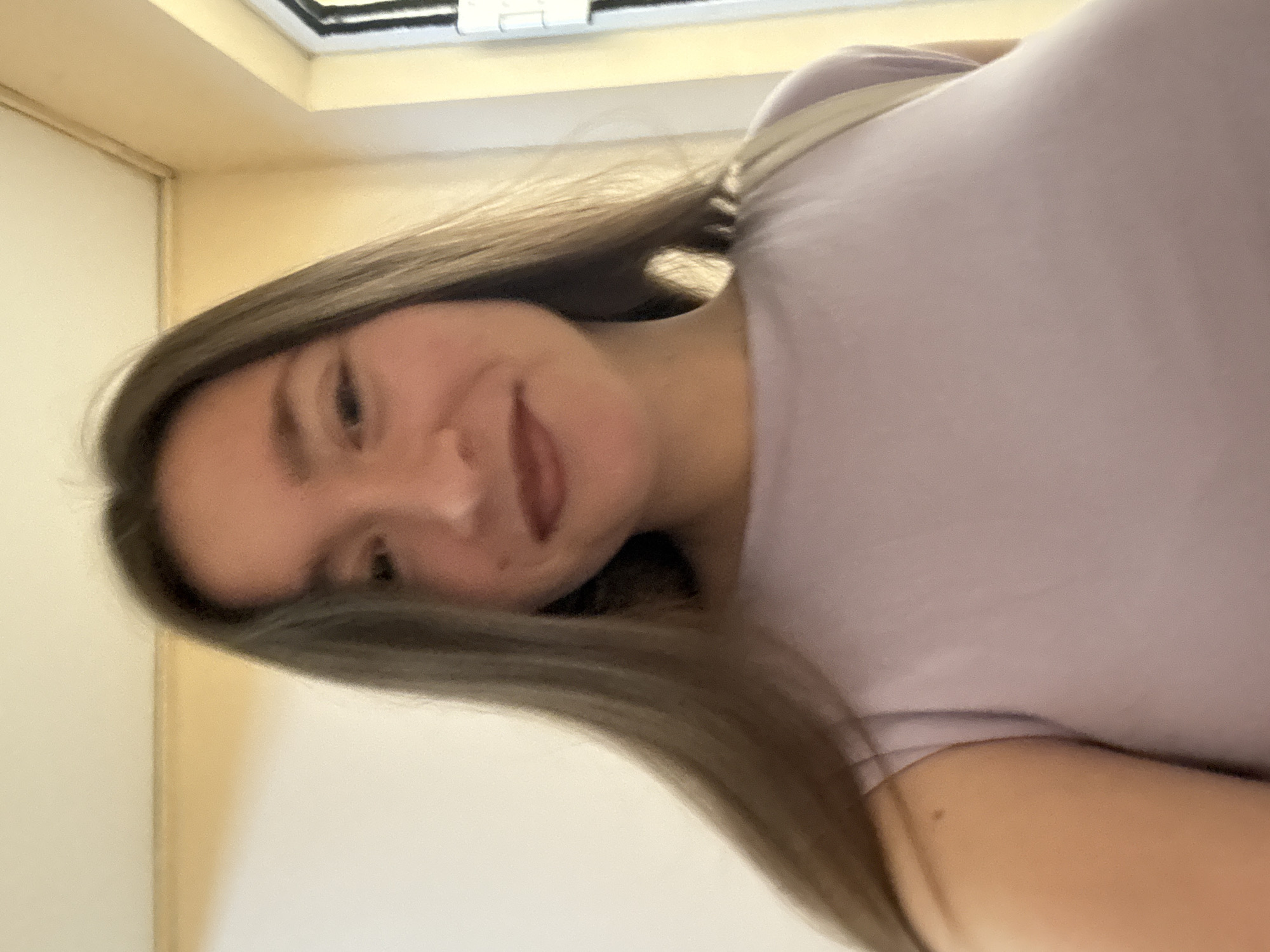 A young woman with long, dark brown hair and fair skin is looking at the camera, smiling slightly. She is wearing a light purple t-shirt and a beige strap is visible across her shoulder. The background consists of a wall with a yellow trim near the ceiling and a window on the right side of the frame. The lighting suggests an indoor setting, possibly a hallway or a room with a window. There are no other people or specific objects that would indicate a precise location within the city of Sofia, Bulgaria.