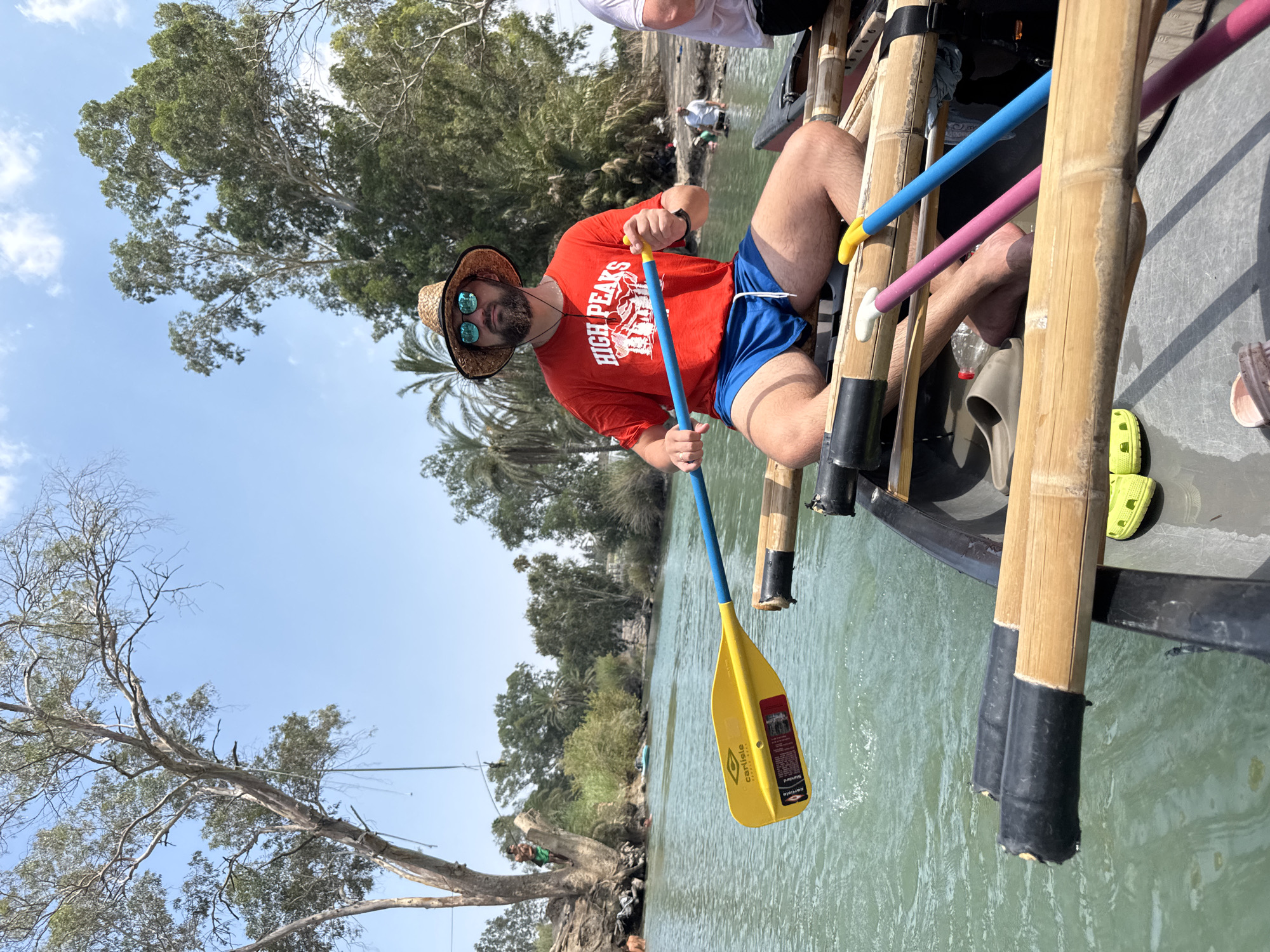A man wearing a straw hat, sunglasses, a red t-shirt with "HIGH PEAKS" printed on it, and blue shorts is seated in a raft, holding a yellow and blue paddle. The raft is on a body of water, likely the Jordan River, in Emek HaYarden Regional Council, Israel, given the context of similar images. The water is a greenish-blue color, and the riverbanks are lined with trees and vegetation. In the background, other people are visible on the river and on the shore. One person is climbing a tree by the riverbank. The sky is clear with a few white clouds. The raft is constructed from bamboo poles and has additional paddles resting within it. The image captures a leisurely outdoor activity.
