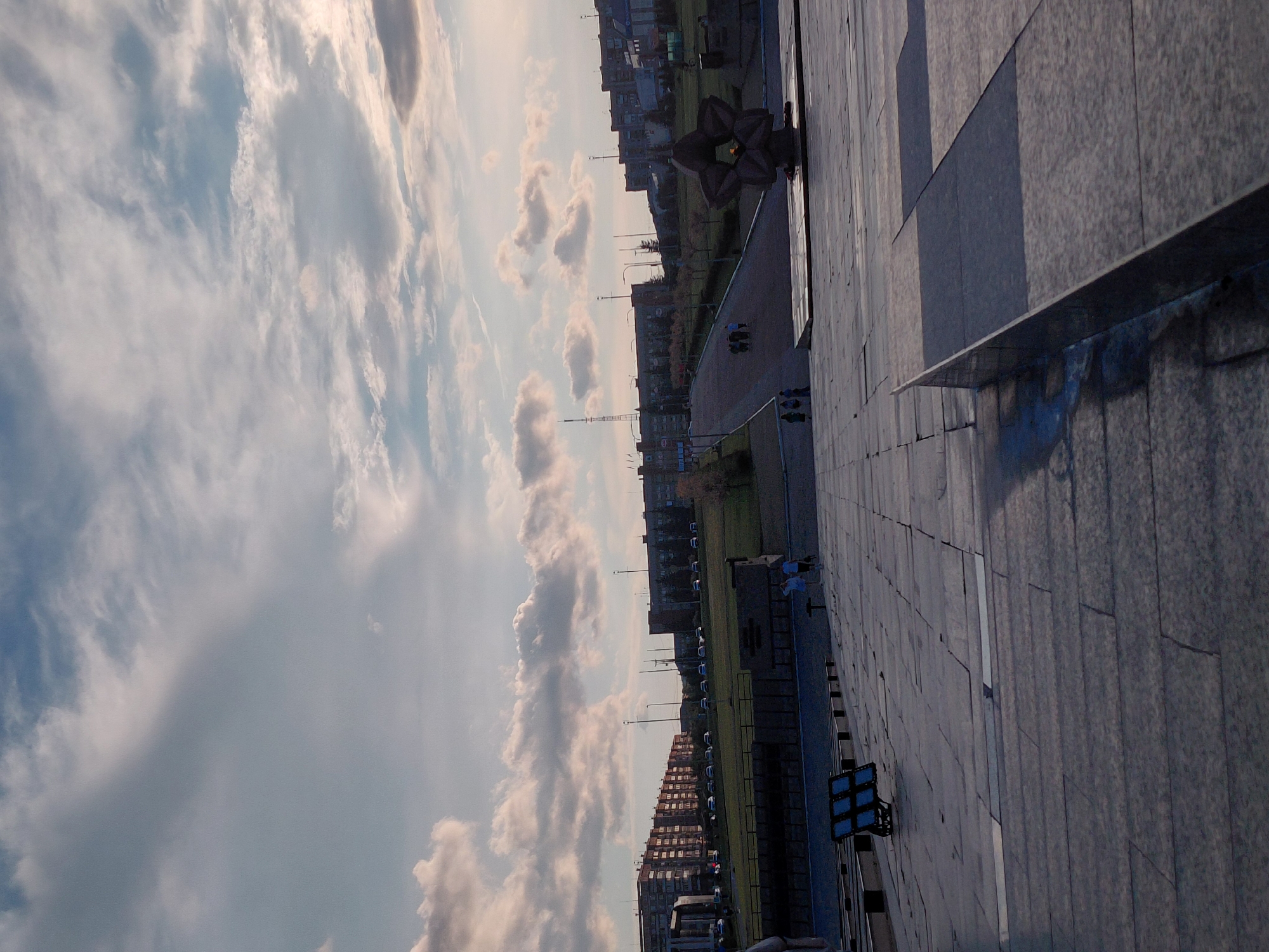 The image depicts a view of a city, likely Magnitogorsk, Russia, from a high vantage point. The scene is dominated by a vast sky filled with dramatic, wispy clouds, indicating it could be late afternoon or early evening, with the sun casting long shadows. To the right, a large, modern building with a tiled facade takes up a significant portion of the frame. Below, a paved walkway leads to a city park area with grass and trees. In the distance, several multi-story buildings are visible, characteristic of urban architecture.
A prominent, dark, sculptural object resembling a stylized flower or star is situated on the walkway, appearing to be a monument or public art installation. Several people are scattered across the walkway and park, suggesting a relaxed, everyday scene. Some are walking, while others appear to be standing or gathered in small groups. A few streetlights and lampposts are visible along the pathway and within the park. On the facade of the building on the right, a modern lighting fixture with blue lights is mounted. The overall impression is of an urban environment at dusk, with a blend of architectural elements and natural sky. No visible text is present in the image.