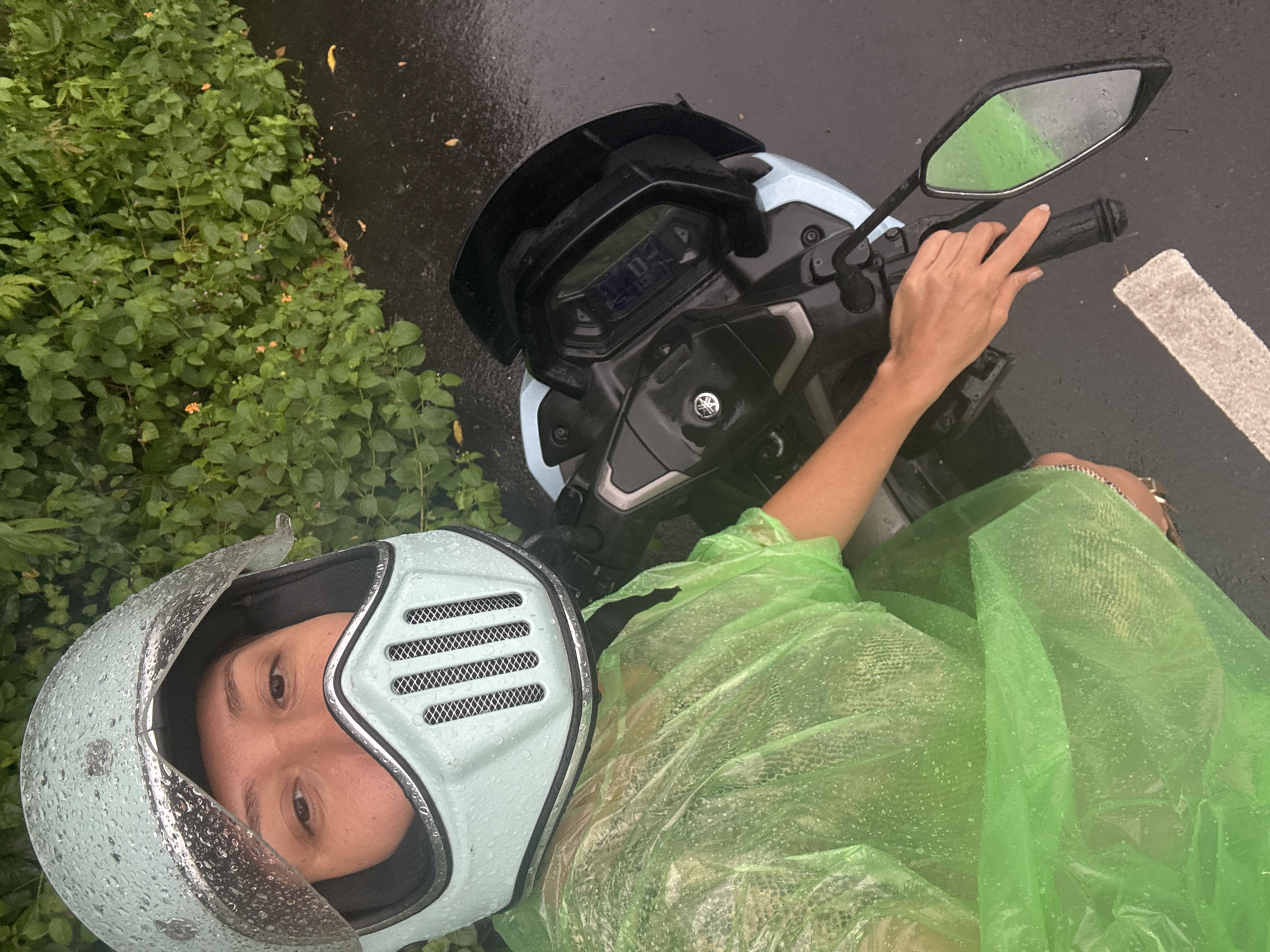 This high-angle shot captures a person on a scooter, wearing a helmet and raincoat, seemingly stopped by a roadside in Kuta Selatan, Indonesia.
The primary subject is an individual, whose face is visible through a light blue helmet adorned with water droplets. The helmet has silver trim and mesh vents. The person wears a transparent, bright green raincoat, also covered in water beads, indicating rain or recent wet conditions. Their right hand rests on the scooter's handlebar.
The scooter itself is light blue and black, with a digital dashboard showing a "0" and other faint indicators, suggesting it might be stationary or at a standstill. A clear "Yamaha" logo is visible near the handlebar. The side mirror reflects the green of the raincoat and possibly the surrounding foliage.
The setting is outdoors, next to a dense patch of lush green bushes, some with small orange flowers. The ground is dark, wet asphalt, implying it is wet from rain. A white road marking is visible on the right side of the frame. The overall lighting appears dim, consistent with an overcast or rainy day. The image has an informal, selfie-like quality due to the direct gaze of the person towards the camera.