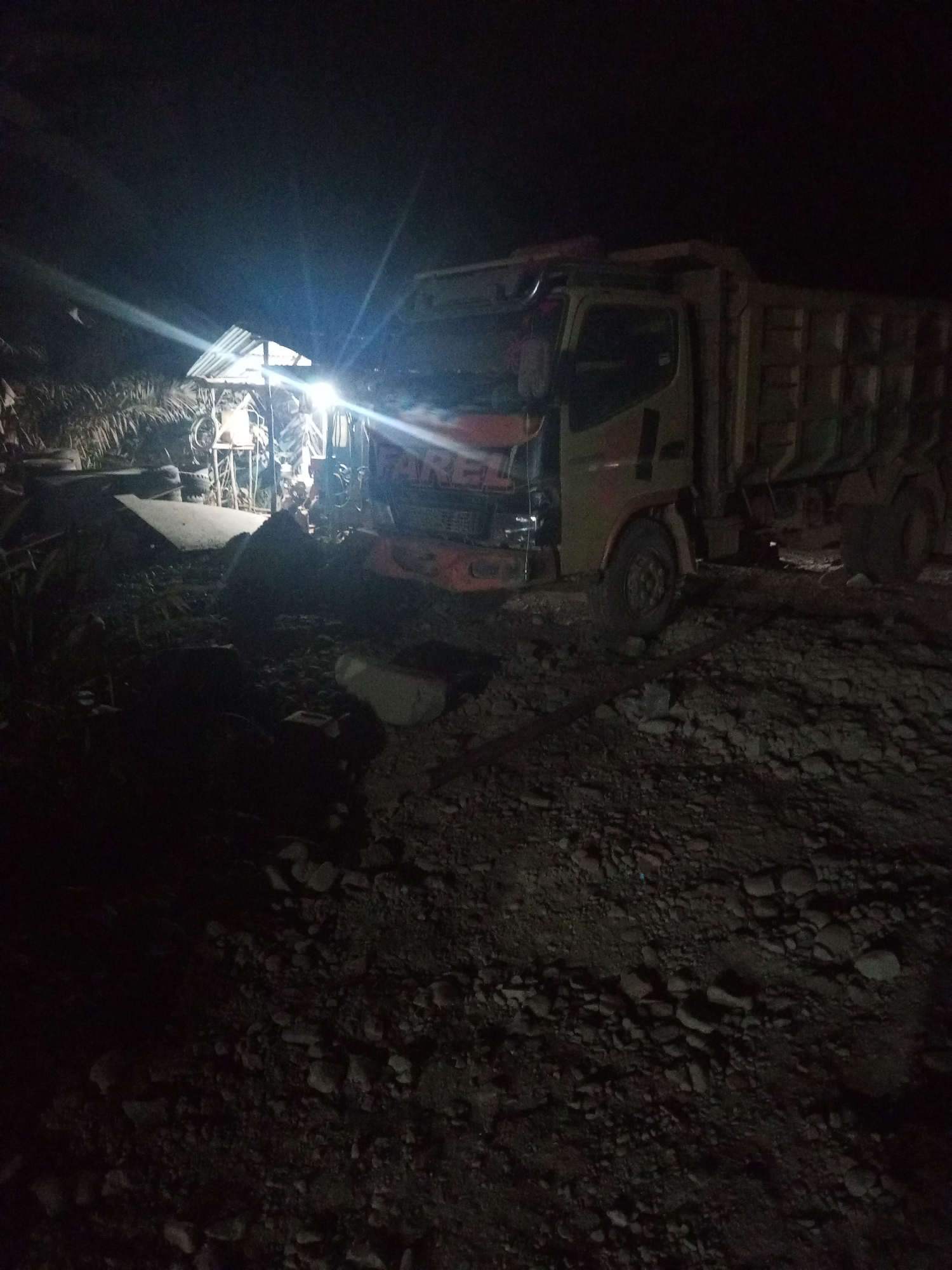 A dump truck is parked on a gravelly area at night. A shed with a corrugated metal roof is situated to the left of the truck, illuminated by a bright light. Palm trees are visible behind the shed. The truck, which appears to be a light yellow or beige color, has "FARE" written on its front grille. The ground is covered in rocks and dirt. The scene is dimly lit, with the bright light from the shed casting harsh shadows. The overall impression is of a rural or industrial area at rest.