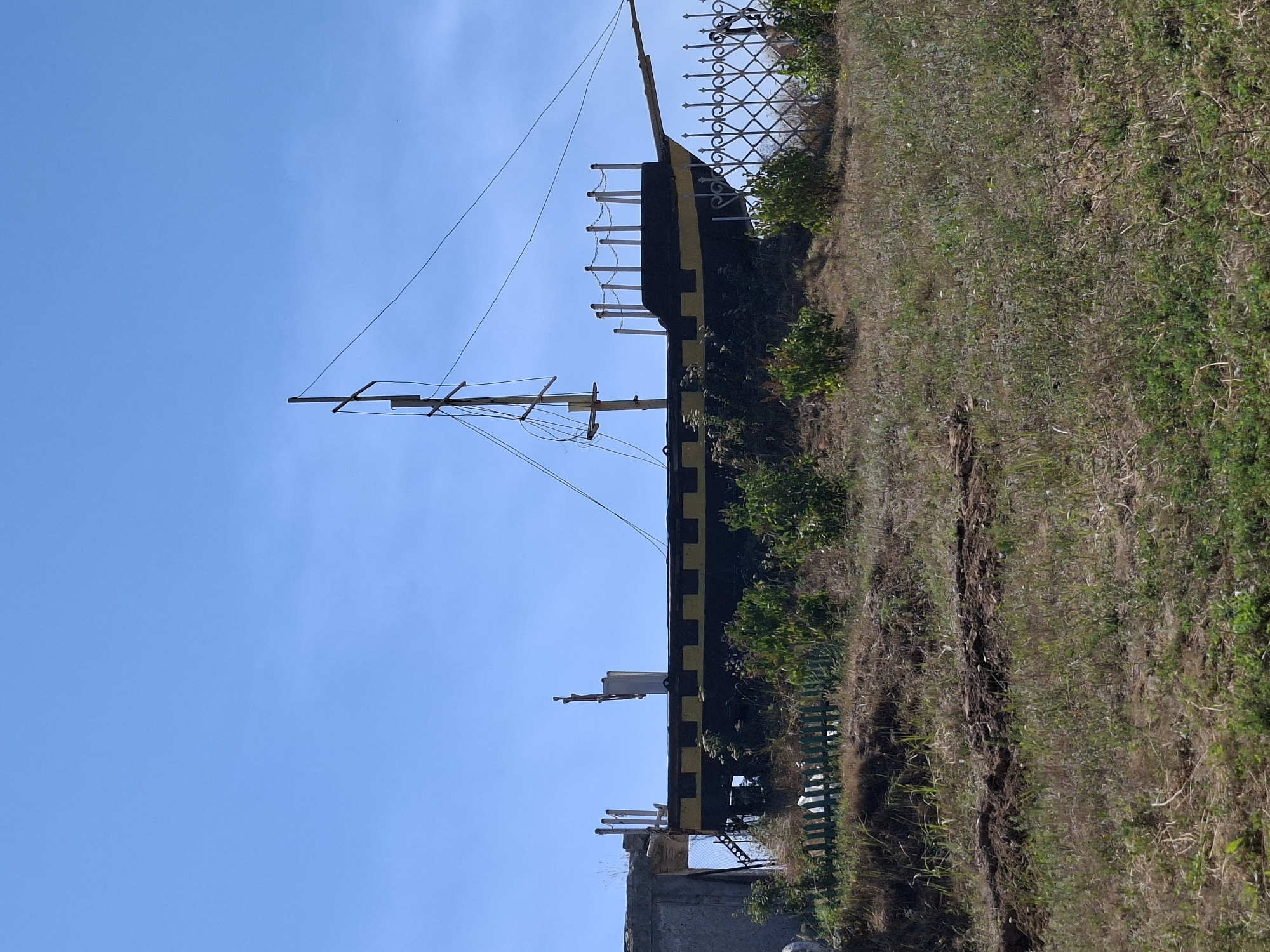 The image shows a dark-hulled, model ship with yellow accents, resting on a grassy hill under a clear blue sky. The ship has a single mast with rigging and a deck railing. In the foreground, there is dry grass, some green bushes, and a short, light green wooden fence. Behind the ship, a decorative metal fence with an ornate pattern is visible. To the left of the ship, a concrete structure partially obscures the view. The scene appears to be an outdoor display or monument.