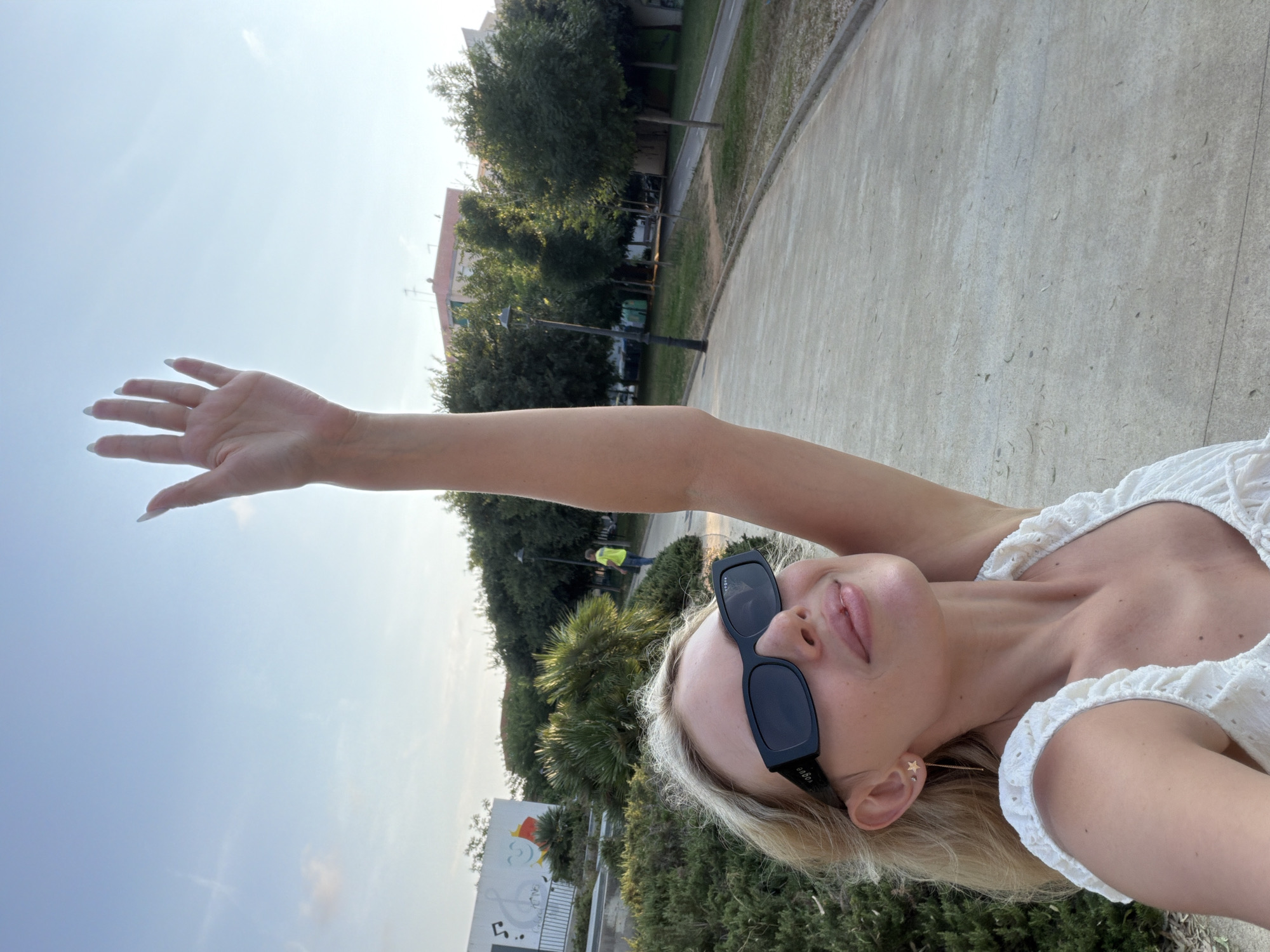 In this selfie, a blonde woman with fair skin and light makeup is shown outdoors. She is wearing a white, eyelet-trimmed top and dark-rimmed sunglasses. A delicate star-shaped earring is visible in her left ear. Her right arm is raised, with her hand extended towards the sky. The setting appears to be a park or public outdoor space with a paved pathway, grass, trees, and some buildings in the background. The sky is clear and blue, suggesting a sunny day. In the distance, two figures can be seen, one wearing a yellow vest, possibly indicating construction or maintenance work. The overall mood seems relaxed and joyful, possibly indicative of a pleasant day spent outdoors. The image appears to have been taken during daylight hours, likely in the afternoon given the lighting. The location context of Benimàmet-beniferri, Spain, is provided.
