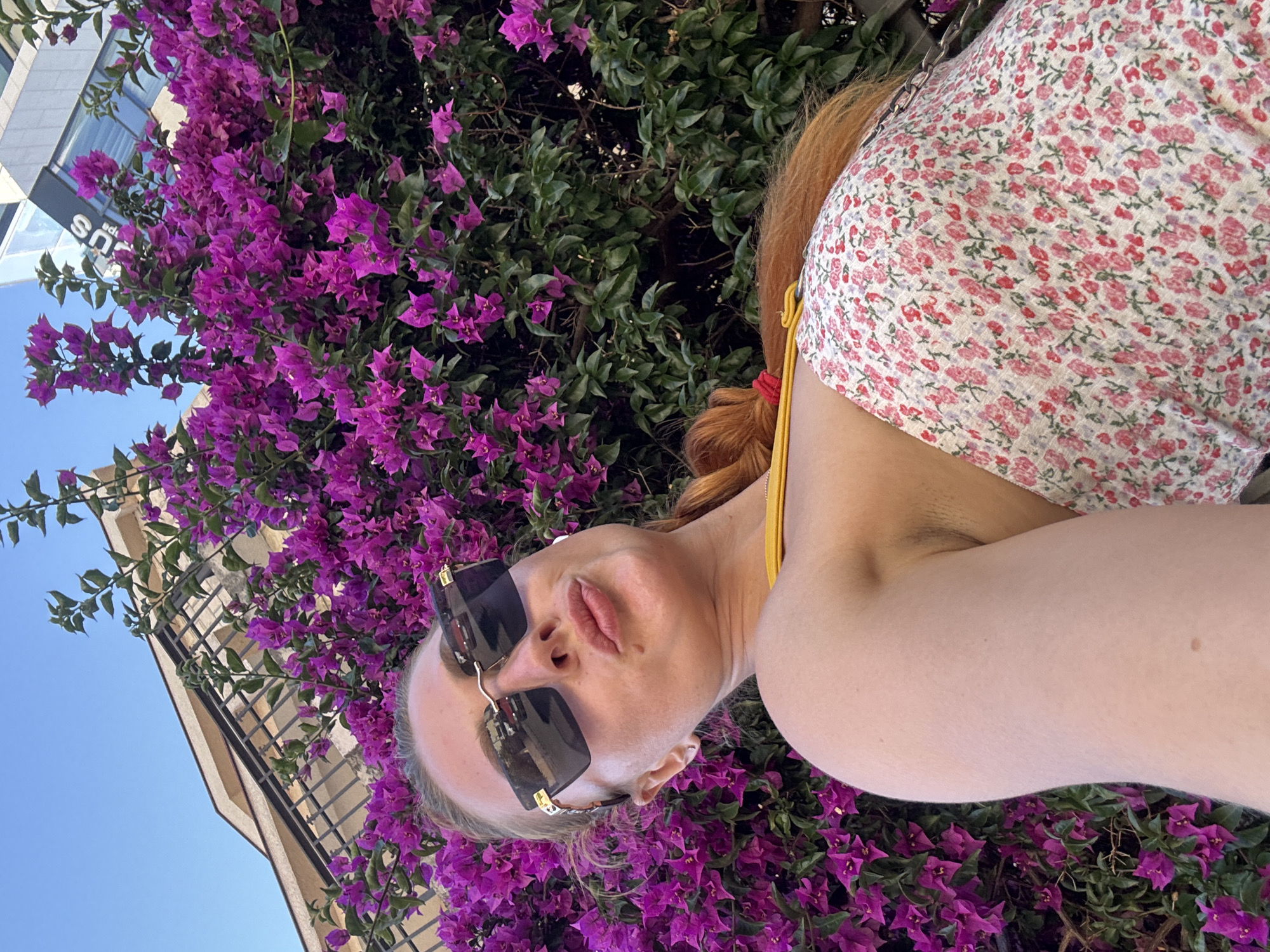 A woman with red hair, wearing large dark sunglasses and a floral-patterned top, poses in front of a vibrant bougainvillea bush in Budva, Montenegro. The bougainvillea is a rich magenta color, with lush green leaves. In the background, parts of a modern building are visible against a clear blue sky. The woman is leaning back, her left arm visible, and her lips are pursed. She has a yellow strap visible on her top, and a small red accessory is tied to her hair. The perspective is a close-up selfie, looking up at the woman.