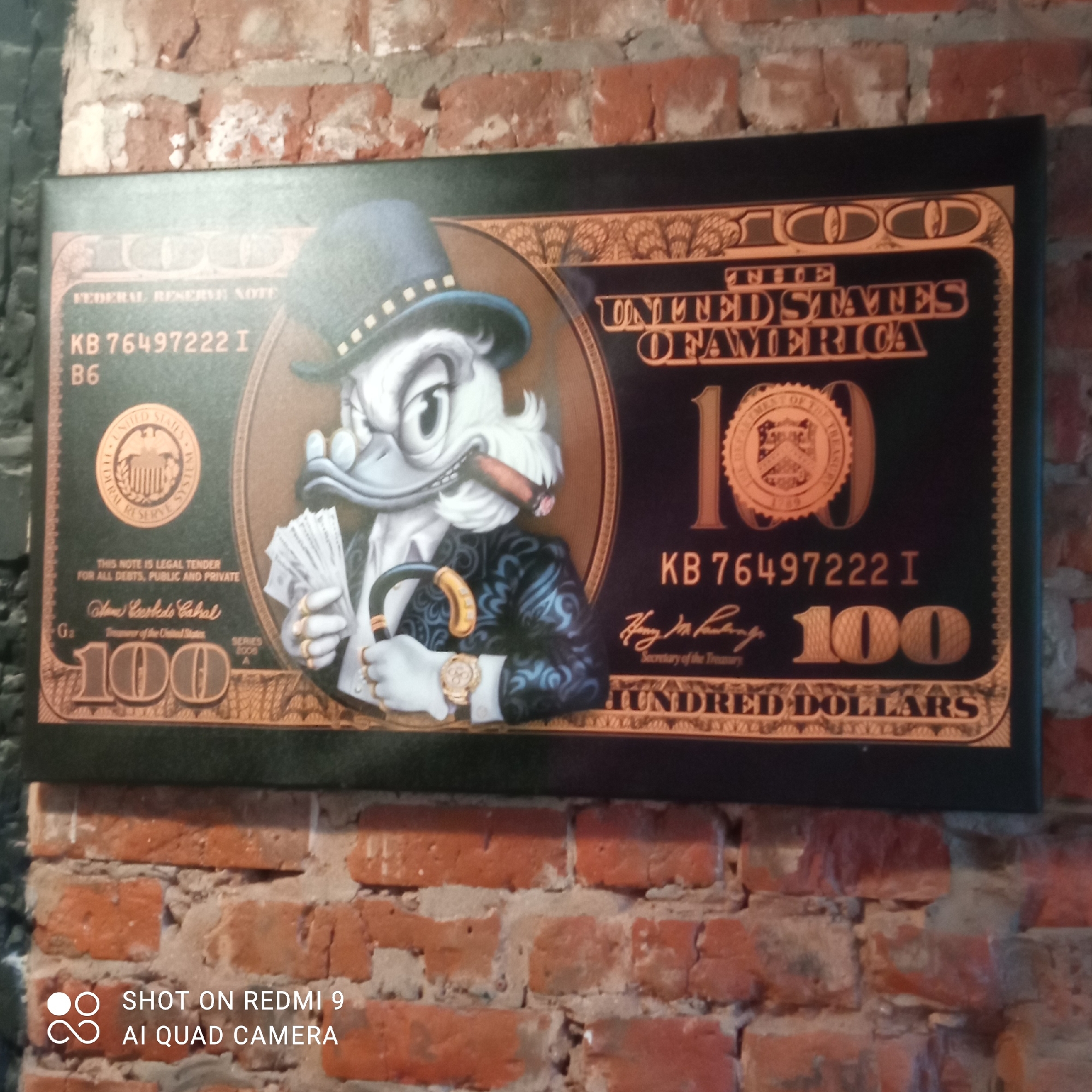 The image depicts a framed print of a stylized hundred-dollar bill featuring Scrooge McDuck. Scrooge is wearing a top hat, a patterned suit, and a wristwatch, holding a cigar and a cane, with a wad of cash in his other hand. The bill's details are altered to reflect the character, with Scrooge's portrait replacing the usual depiction of Benjamin Franklin. The print is mounted on a brick wall. The photo was taken by a Redmi 9 AI Quad Camera and is located in the city of Chaikovsky, Russia.