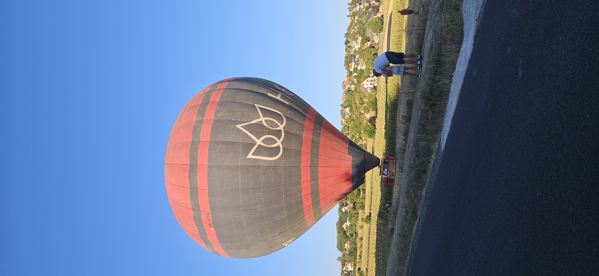 A hot air balloon, predominantly dark gray with red horizontal stripes and a white floral logo, is partially inflated and tethered to a wicker basket on the ground. The balloon is positioned in a field, likely a sunflower field given the yellow blooms visible. In the foreground, a paved road runs diagonally across the bottom of the frame. Behind the balloon, a village with numerous houses, nestled on a hillside covered in trees, is visible under a clear blue sky. Two individuals, a man and a child, stand on the grassy verge near the road, facing the balloon. Another child in a red shirt is seen running in the background to the right. The scene appears to be set in Cserszegtomaj, Hungary, during what seems to be an early morning or late afternoon, judging by the lighting and clear sky.