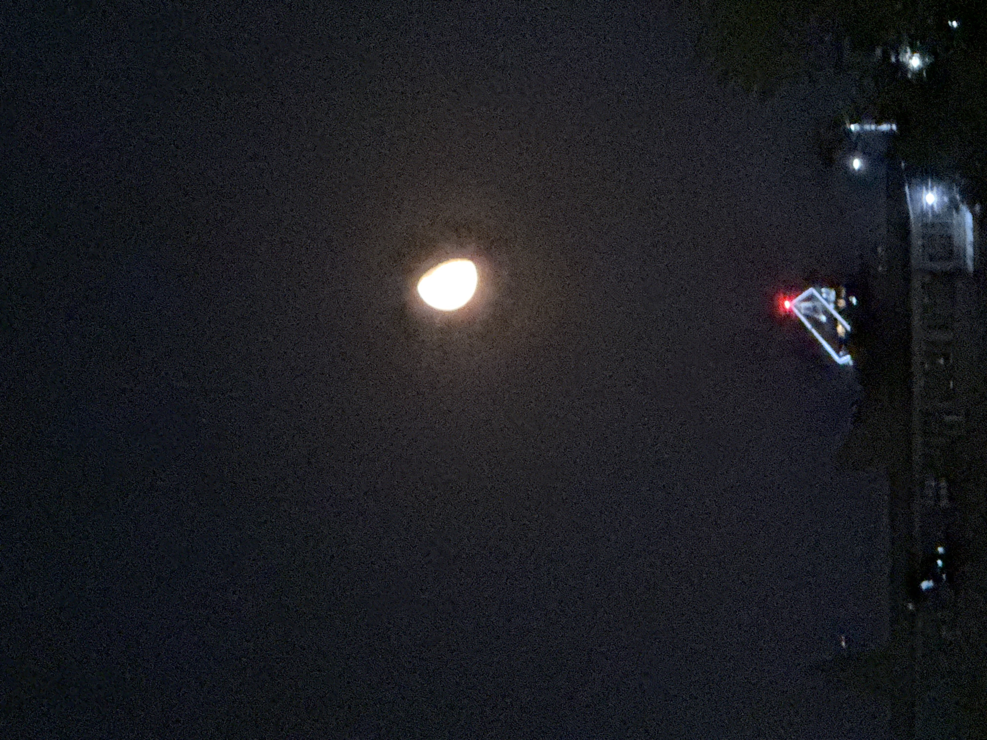 A night photograph captures a crescent moon in the upper right quadrant of the frame. Below the moon, a triangular structure with bright white and red lights is visible, likely a building or antenna. The lower portion of the image shows silhouetted rooftops and trees, with various lights suggesting an urban environment. The sky is dark with a visible grain, indicating a low-light photograph. The scene appears to be set in a city at night.