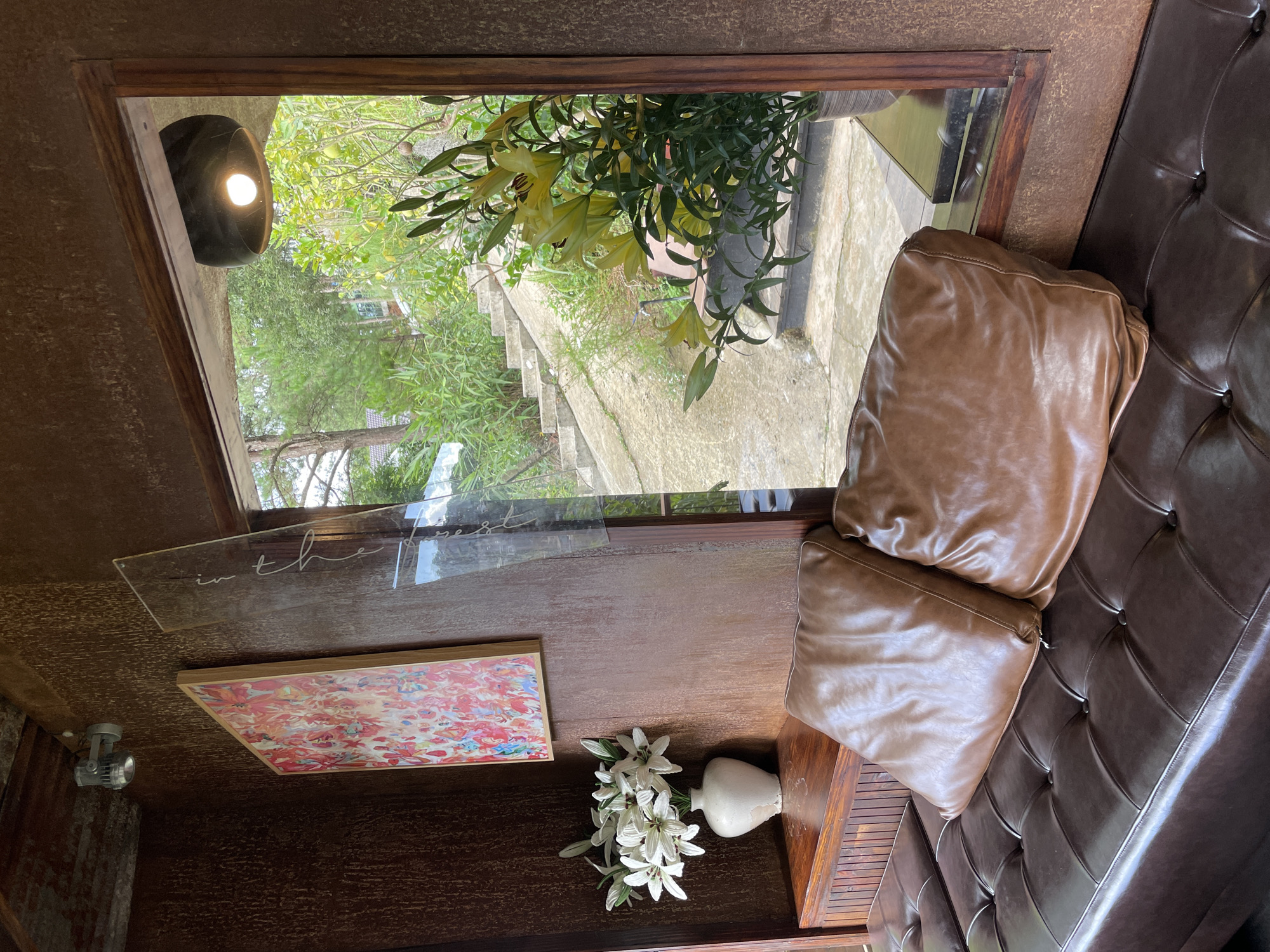 The image depicts an interior scene looking out through a window onto an outdoor staircase and lush greenery. The indoor space features a dark brown, tufted leather couch with two brown leather cushions on it. To the left, a wooden shelf holds a white vase with white flowers and a framed abstract painting with red and pink hues on the wall. A light fixture is visible on the upper left wall.
Through the window, a concrete staircase with greenery on either side leads upwards. In the foreground outside the window, a potted plant with yellow lilies is visible. The overall aesthetic suggests a cozy and rustic ambiance, possibly a cafe or a private residence. The location is identified as Da Lat, Vietnam.