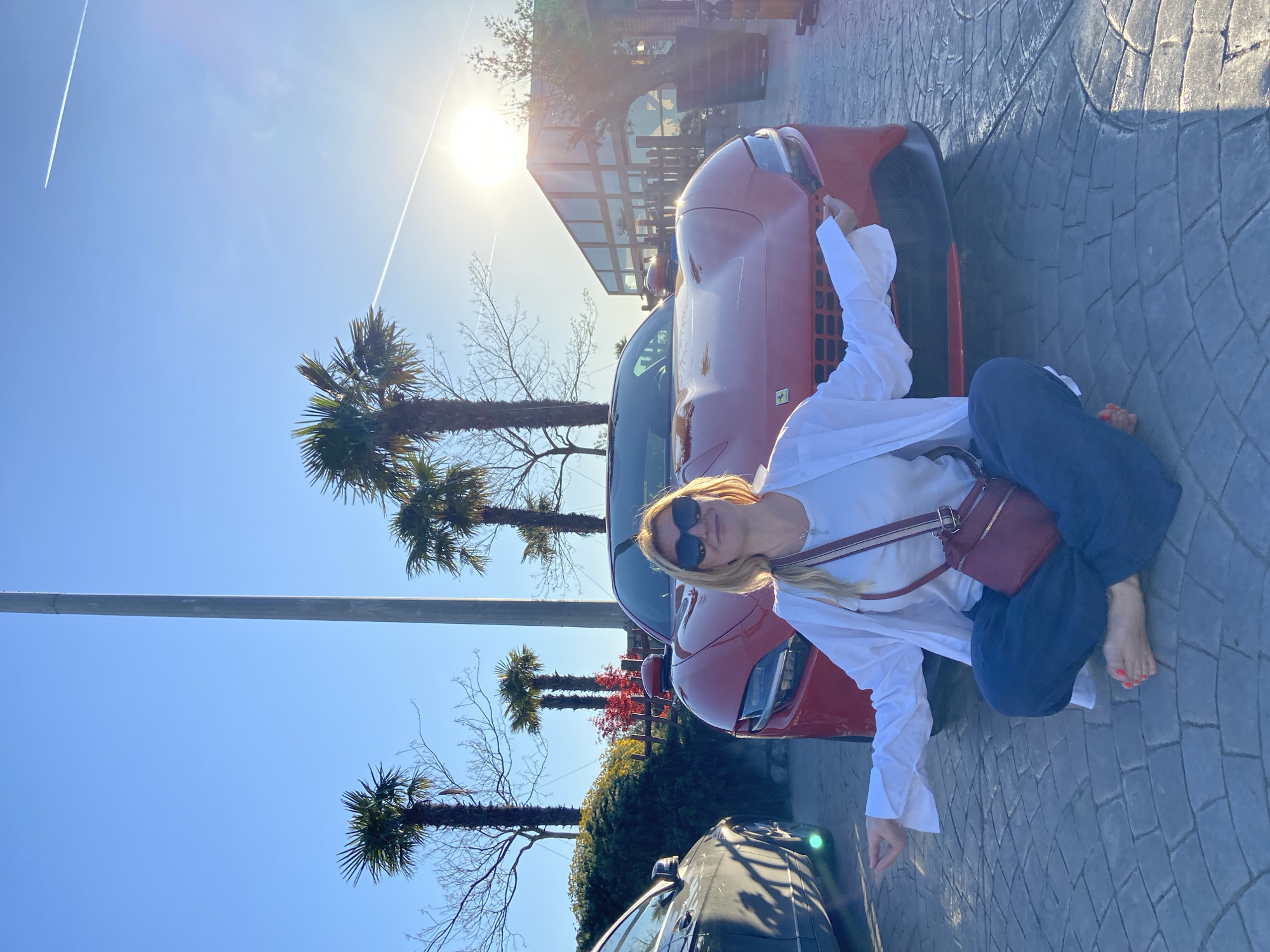 A woman is squatting barefoot on a paved surface next to a red car under a sunny sky, likely in Kovačko Polje, Montenegro. She wears sunglasses, a white top, an unbuttoned white shirt, and blue pants. A maroon purse hangs across her chest. Palm trees and a modern glass building are visible in the background. A dark car is parked behind the red one. The sun is bright, creating lens flare and a contrail can be seen in the blue sky.