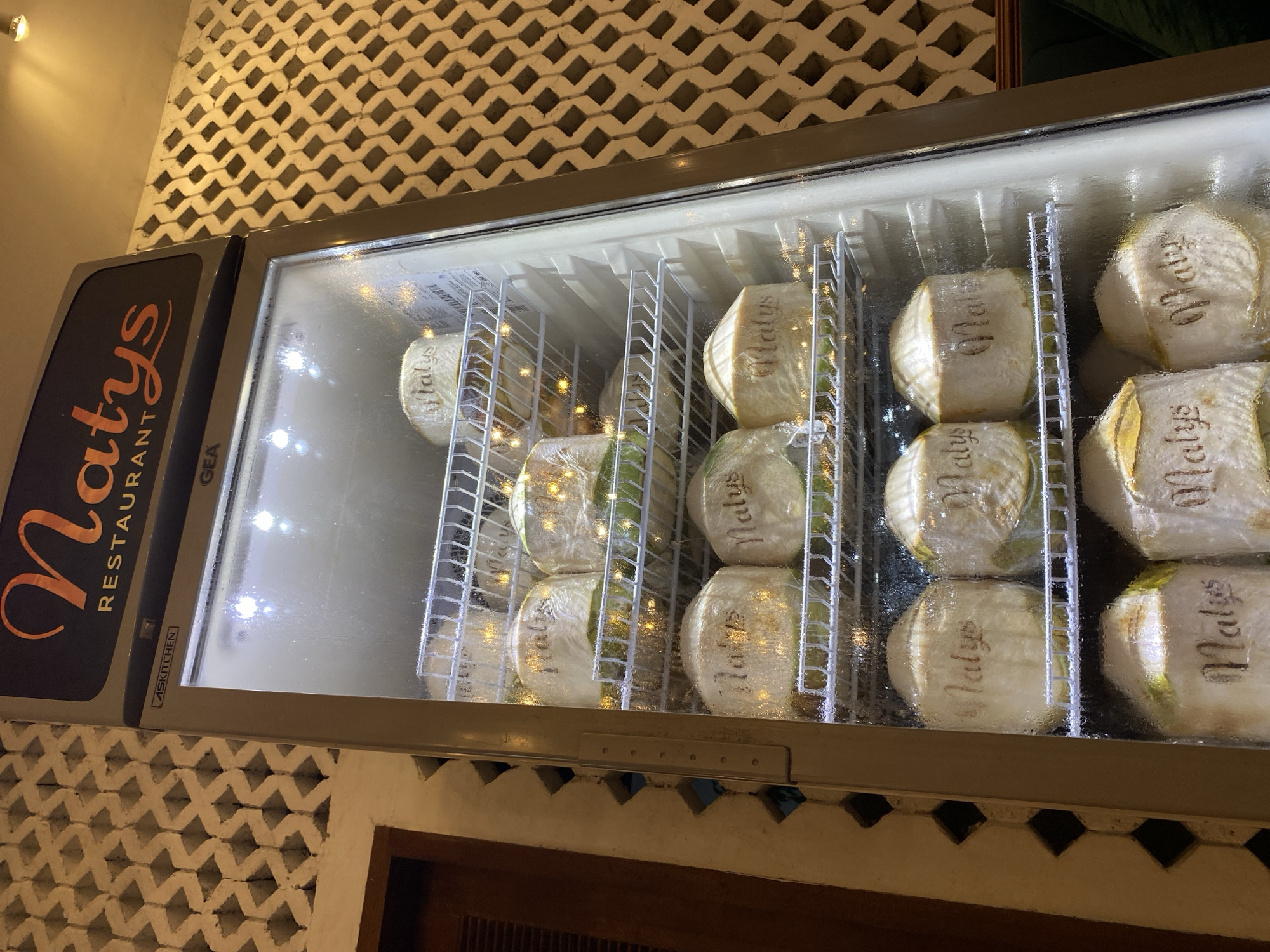 A refrigerated display case at Natys Restaurant in Nusa Dua, Indonesia, showcases neatly stacked, peeled coconuts wrapped in plastic. The coconuts feature the restaurant's name, "Natys," printed on their surfaces. The display case, with its metallic frame, is situated against a textured wall with a unique geometric pattern. The ambient lighting and condensation on the display case's glass front indicate a cool, humid environment. The shelving within the display case is wire-framed, allowing for optimal air circulation around the produce. The overall scene depicts a controlled storage of fresh coconuts, ready for service at the restaurant.
