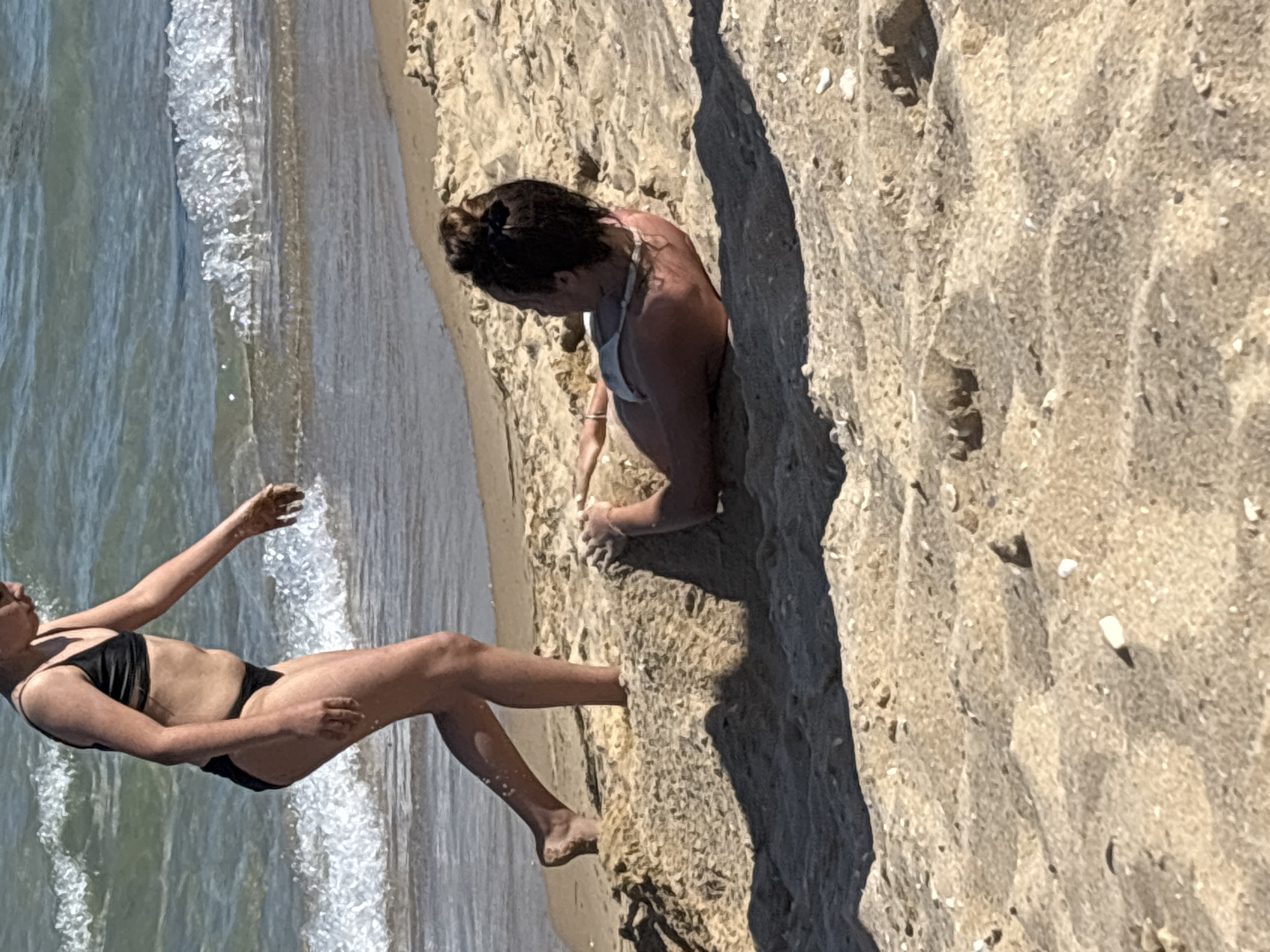 Two individuals are on a sandy beach near the ocean. In the foreground, a person is partially buried in the sand, lying on their back with their upper body exposed. Their head is turned to the side, and their arms are resting on the sand. They are wearing a light-colored bikini top. In the background, another person is standing in shallow water at the edge of the beach. They are wearing a dark bikini and are facing away from the viewer, with their right leg extended as if stepping onto the sand. The ocean water is blue and appears to have small waves. The sand is light brown and textured, with some shell fragments visible. The lighting suggests it is a sunny day.