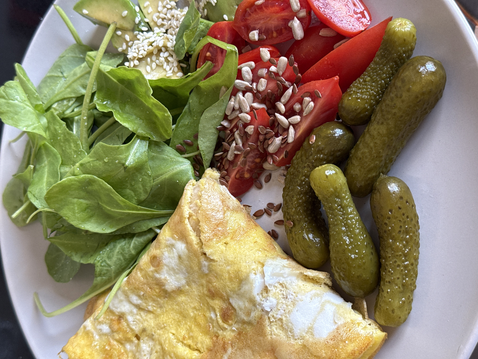 A close-up shot depicts a plate of food consisting of an omelet, mixed greens, sliced tomatoes, avocado, pickles, and various seeds, possibly taken in Tel Aviv, Israel. The omelet is light yellow with cooked white egg visible. The greens appear fresh and slightly moist, with a mix of spinach. The tomatoes are cut into wedges and sprinkled with sunflower seeds and flax seeds. The avocado slices are topped with sesame seeds. Several small pickles are arranged on the right side of the plate. The plate is light-colored. The background is blurred, suggesting a shallow depth of field.