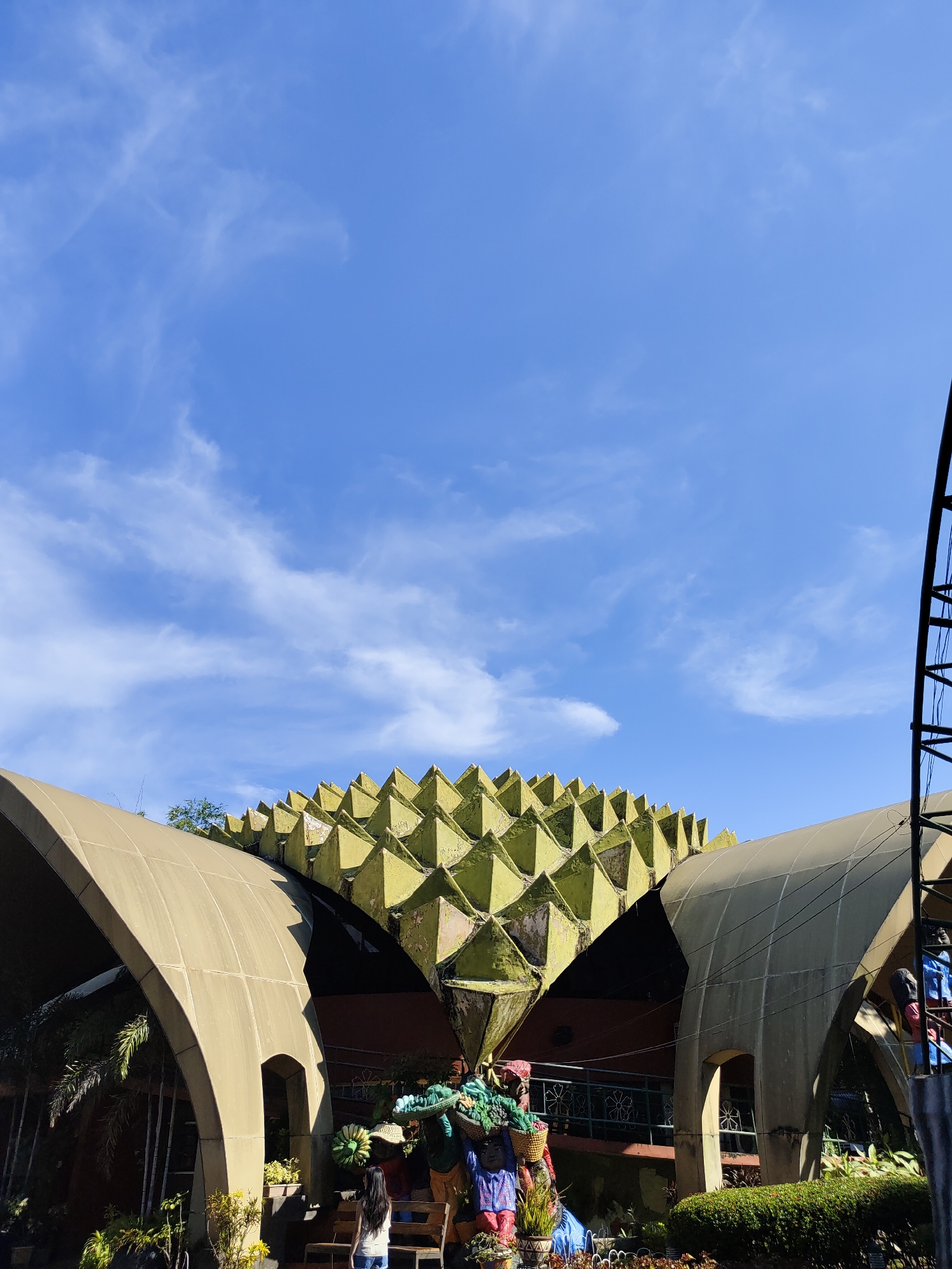 This is a photograph of the Durian Garden in Davao City, Philippines, taken on a clear day with a bright blue sky and scattered white clouds. The focal point of the image is a unique, durian-shaped building with a spiky green roof and curved beige overhangs. Several colorful statues depicting people carrying baskets of what appear to be fruits and vegetables are arranged in front of the building, adding a vibrant touch to the scene. A young woman with long black hair is standing near the statues, facing away from the camera. Lush greenery, including potted plants and bushes, surrounds the statues and the base of the building, suggesting a garden setting. The overall impression is one of a quirky, artistic, and tropical tourist attraction.