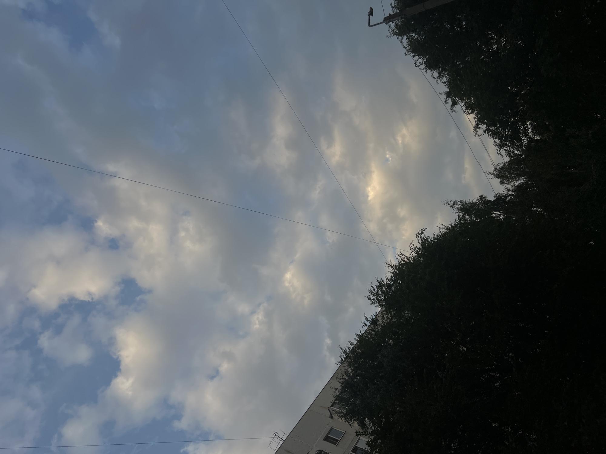 The image shows a view of the sky with clouds above a partially visible building and trees. The building appears to be a multi-story residential structure with windows. Dark green trees line the bottom of the frame, obscuring much of the ground level. Two thin wires stretch across the sky, forming diagonal lines. A street lamp is visible on the right side, near the top of the frame. The sky is a mix of blue and gray clouds, with some lighter patches suggesting sunlight. The overall impression is an urban landscape, likely taken during daylight hours with overcast or partly cloudy conditions. The specific location in Ulyanovsk, Russia is not discernible from the visual elements alone.