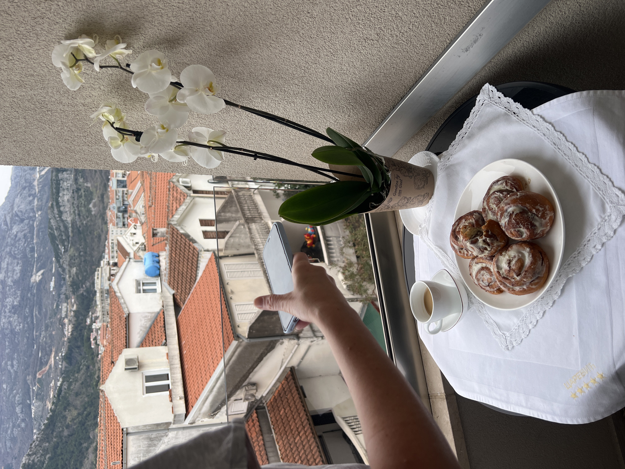 The image shows a person taking a photograph with a smartphone on a balcony in Budva, Montenegro. The balcony overlooks a cityscape with red-tiled roofs and mountains in the background. On the balcony table, covered with a white lace tablecloth, is a plate of pastries, a cup of coffee, and a potted orchid. The person's arm and hand are visible as they hold the smartphone. The lighting suggests it is daytime, and the scene appears peaceful and leisurely.