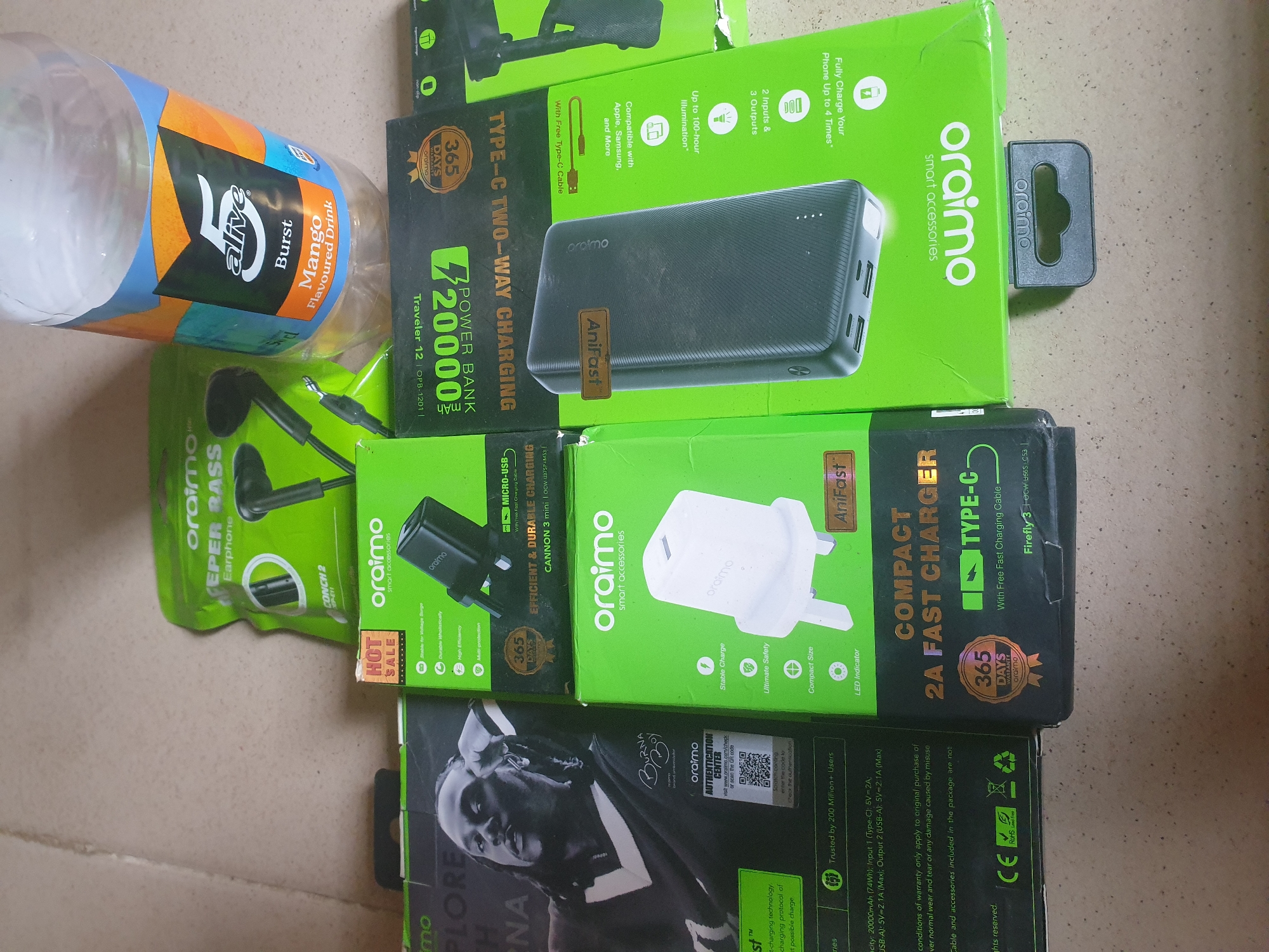 The image depicts a collection of electronic accessories and a partially consumed bottle of "5 alive Burst Mango Flavoured Drink" on a textured surface, likely a tabletop or floor. The items are predominantly Oraimo branded, including a green package of "EPER RASS Earphone," a green box for a "20000mAh POWER BANK" with "TYPE-C TWO-WAY CHARGING" capabilities, and a white "2A FAST CHARGER" in a green box. Another Oraimo product, a black power bank, is also visible within its green packaging. A black and white image of a person with dreadlocks, potentially an advertisement for a music artist, is partially visible on the left side of the frame. The background is out of focus, but suggests an indoor setting. The overall impression is that of consumer electronics and beverages. The reference to "Anilelerin, Nigeria" appears to be an erroneous association as there is no visual information within the image to support this specific location.