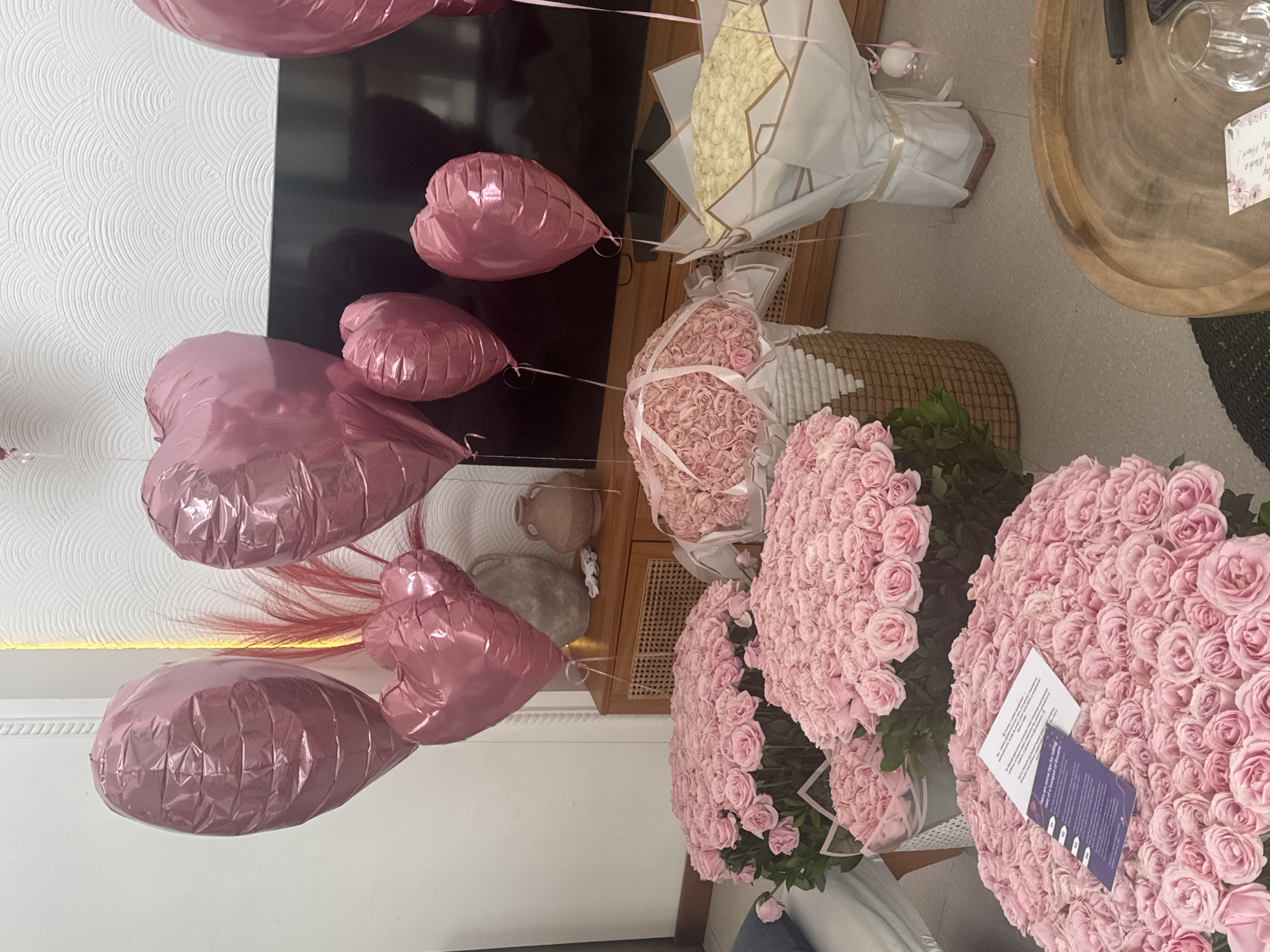 The image shows a room filled with floral arrangements and heart-shaped balloons. Two large bouquets of pink roses are prominently displayed, along with a smaller bouquet of light-colored flowers wrapped in white and gold paper. Several pink heart-shaped balloons float in the air. A wooden coffee table, a wicker basket, and a television can also be seen in the background. The location is North Kuta, Indonesia.