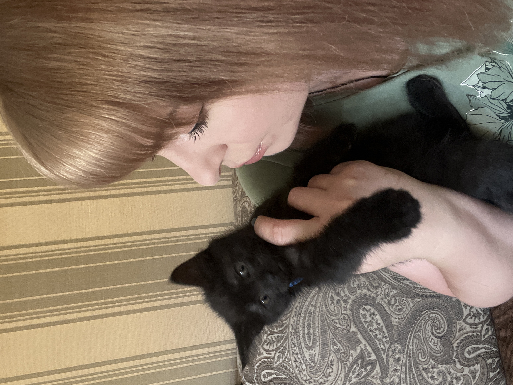 A medium shot captures a woman with light brown hair looking down at a black kitten she is holding. The kitten is nestled in the crook of the woman's arm, its face turned towards the camera. The woman is wearing a green top. The background consists of a striped wallpaper. The scene appears to be indoors, possibly in a residential setting. The image does not provide any information about the city of Shymkent, Kazakhstan.