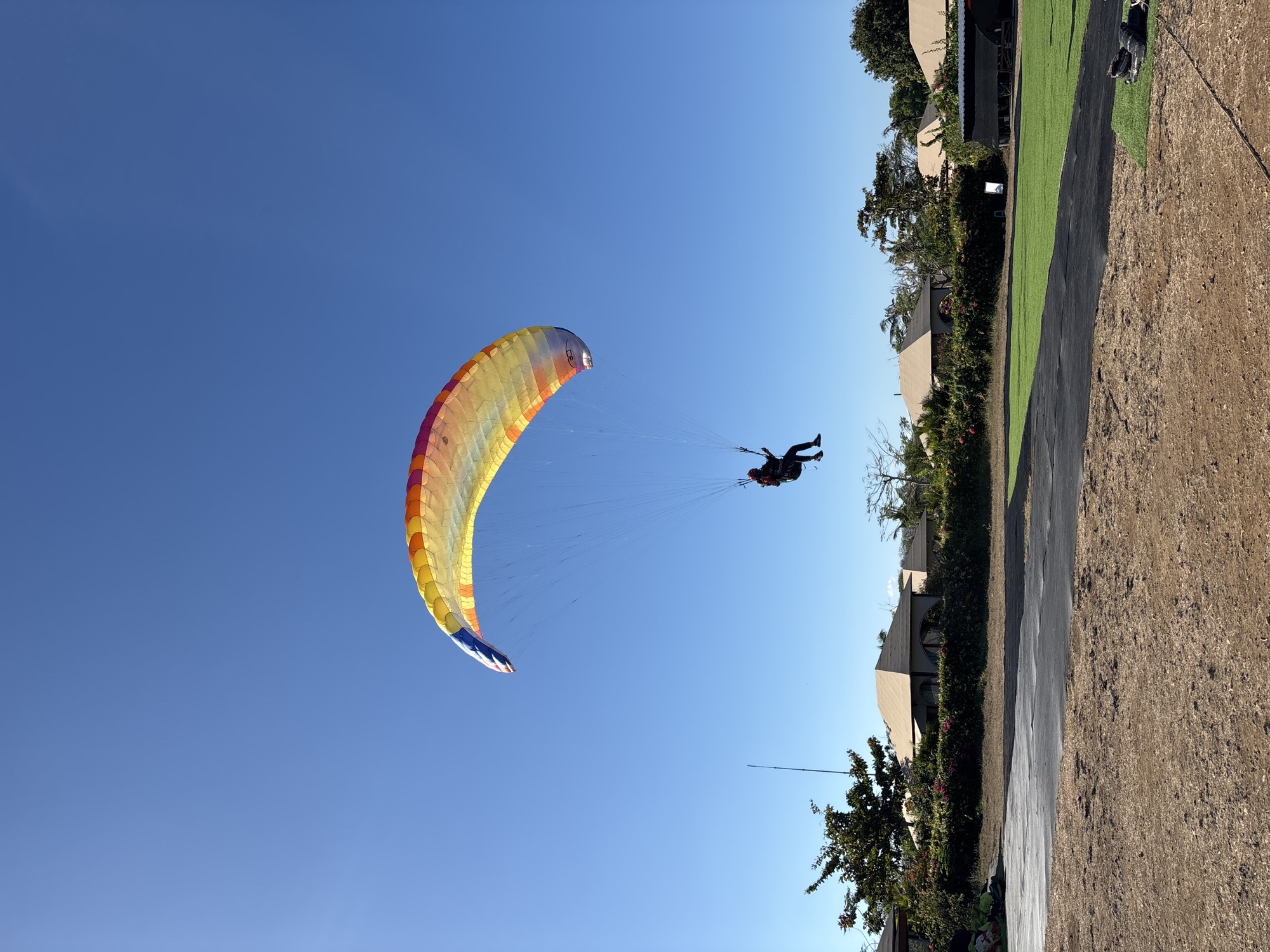 A lone paraglider with a brightly colored wing soars against a clear blue sky over a landscape in Kuta Selatan, Indonesia. The paraglider, wearing a dark suit and helmet, is suspended beneath a rainbow-hued wing, a spectacle of yellow, orange, red, and blue. Below the paraglider, a row of beige, pyramid-roofed buildings are nestled behind a dense hedge of green foliage and vibrant pink flowers. In the foreground, a patch of well-maintained green grass is bordered by an area of dry, brown earth. The scene evokes a sense of freedom and adventure, set against a backdrop of tropical architecture and natural beauty.
