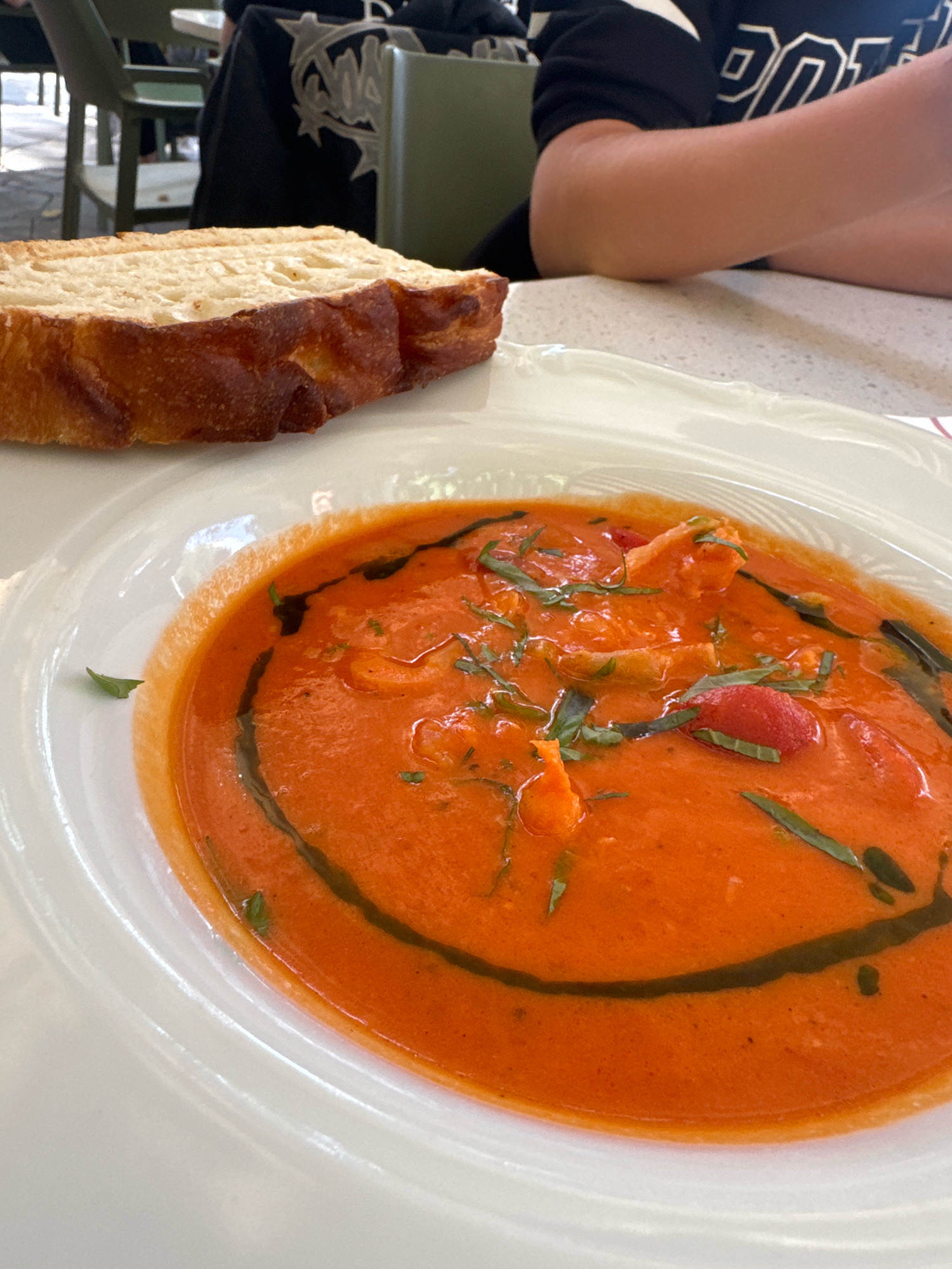 A close-up, eye-level shot shows a white bowl of creamy tomato soup with garnishes of basil and what appears to be cherry tomatoes, swirled with a dark oil. To the left of the bowl, a thick slice of toasted bread rests on the rim of the bowl. The setting appears to be an outdoor cafe or restaurant with a partially visible person in the background wearing a black top with white lettering. The person's arm is resting on a white speckled table. The overall scene is brightly lit, suggesting daytime. The image does not contain any information that can be used to identify its location in Almaty, Kazakhstan.