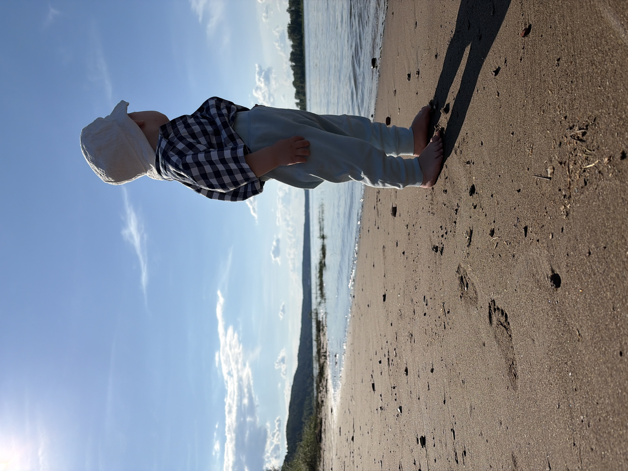 A young child wearing a white hat, a checkered shirt, and light blue pants stands on a sandy beach. The child is facing away from the camera, looking towards a body of water. The water is calm and reflects the blue sky, which is dotted with white clouds. In the distance, a tree-covered hill or embankment forms the horizon. The foreground consists of wet sand with footprints. The overall scene appears to be a sunny day at the Olyakhovka, Russia waterfront.