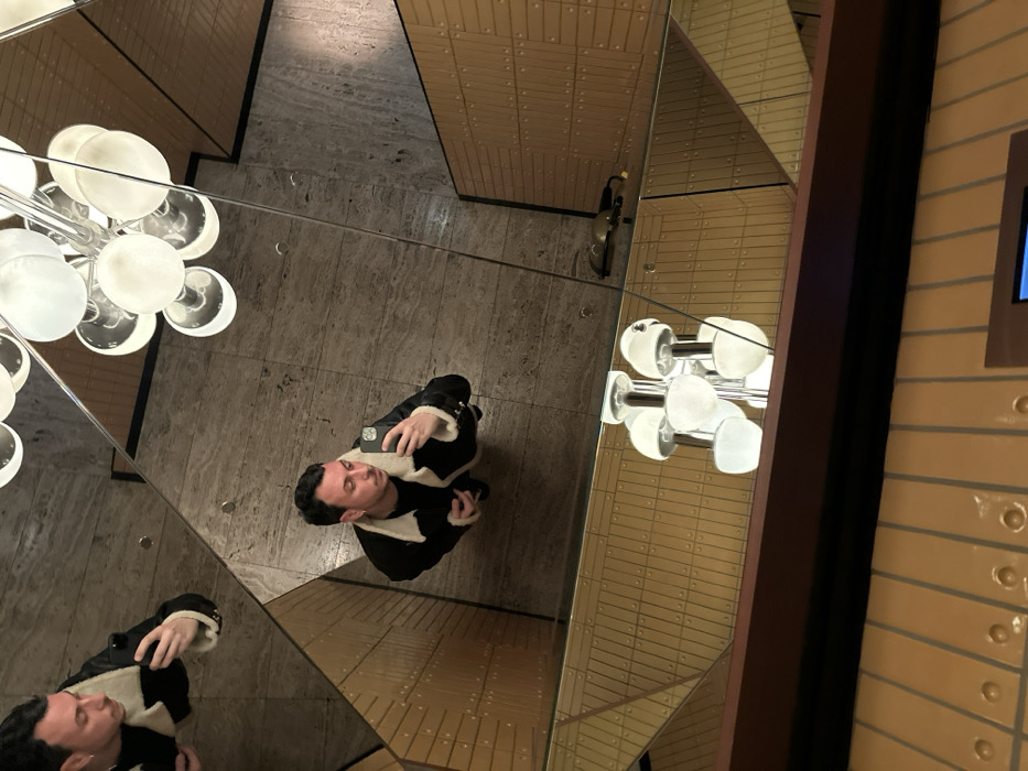 A person wearing a black jacket with white trim is standing in a geometrically mirrored space. The individual is holding a phone, seemingly taking a selfie. Above and to the left, a chandelier-like light fixture is visible. The surrounding walls and floors are covered in a pattern of reflective surfaces, creating multiple mirrored images of the person and the environment. The color palette is dominated by neutral tones and highlights, with a mix of metallic elements.
