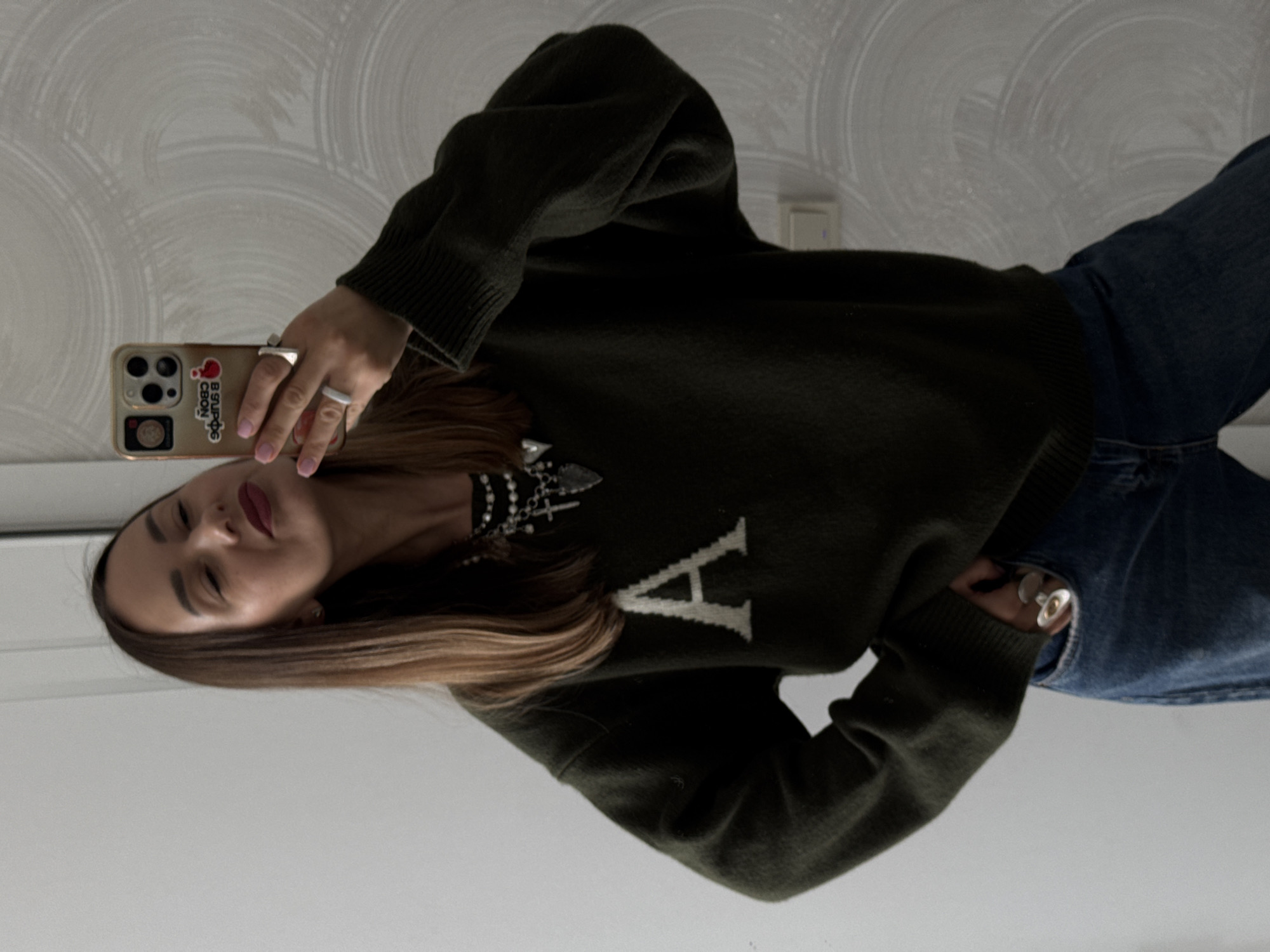 A woman is taking a selfie in a mirror. She is wearing a dark green sweater with a large white "A" on the front, blue jeans, and a necklace. She has long brown hair and is wearing red lipstick. She is holding a tan iPhone with stickers on the back. The background is a wall with a patterned wallpaper. There is no indication that this is in Sochi, Russia.
