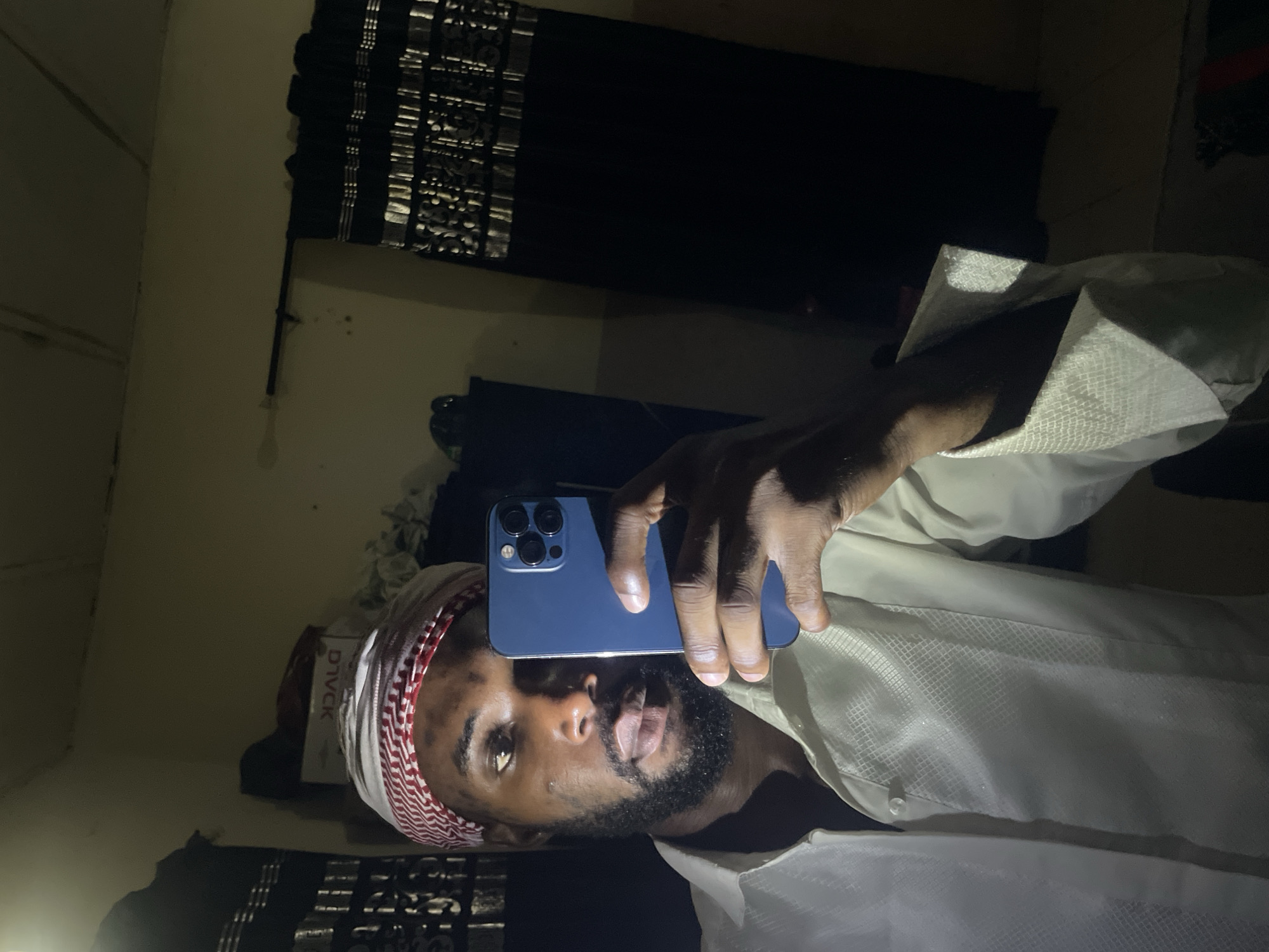 A man with brown skin, a beard, and wearing a red and white patterned head covering is holding a blue smartphone up to take a selfie. He is also wearing a white shirt. The background is dark and appears to be an interior space with patterned curtains or blinds in the background. The location is not specified or identifiable from the image.