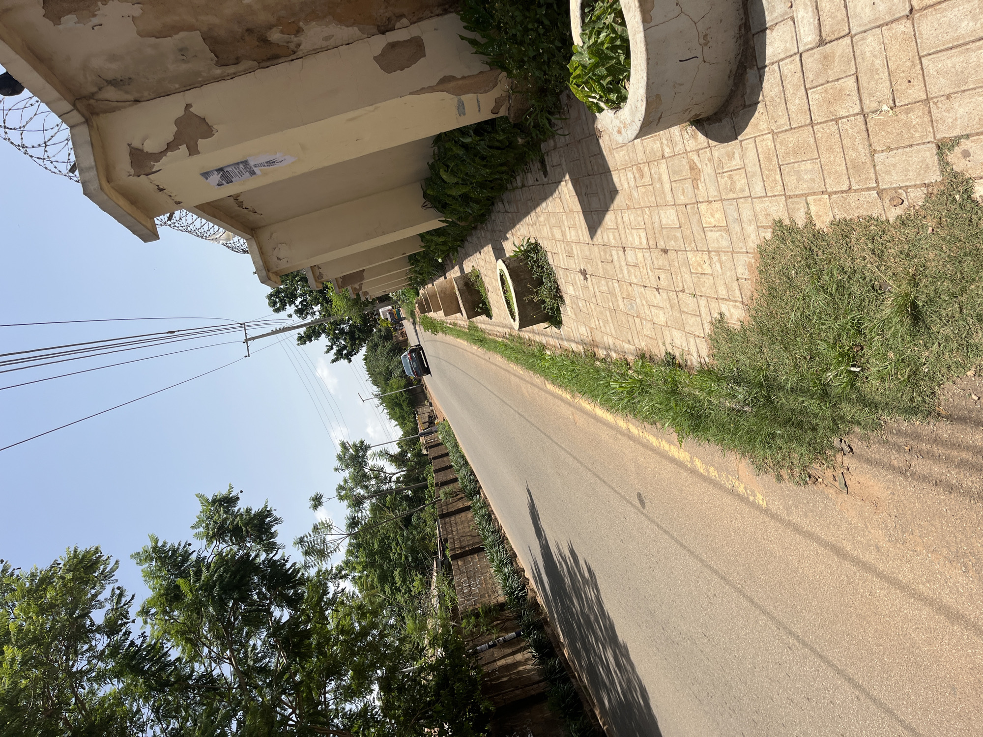 The image shows a street scene in Gura Top, Nigeria. A paved road runs from the foreground into the distance, bordered on the left by a brick wall and trees, and on the right by a sidewalk with a decorative wall and planters. A dark-colored sedan is visible driving away from the camera on the road. Overhead power lines are strung across the sky. The wall on the right is light beige with peeling paint and topped with barbed wire. The sidewalk is made of interlocking pavers and several circular concrete planters containing greenery are spaced along its edge. Lush green grass grows along the side of the road. The sky is clear and blue.