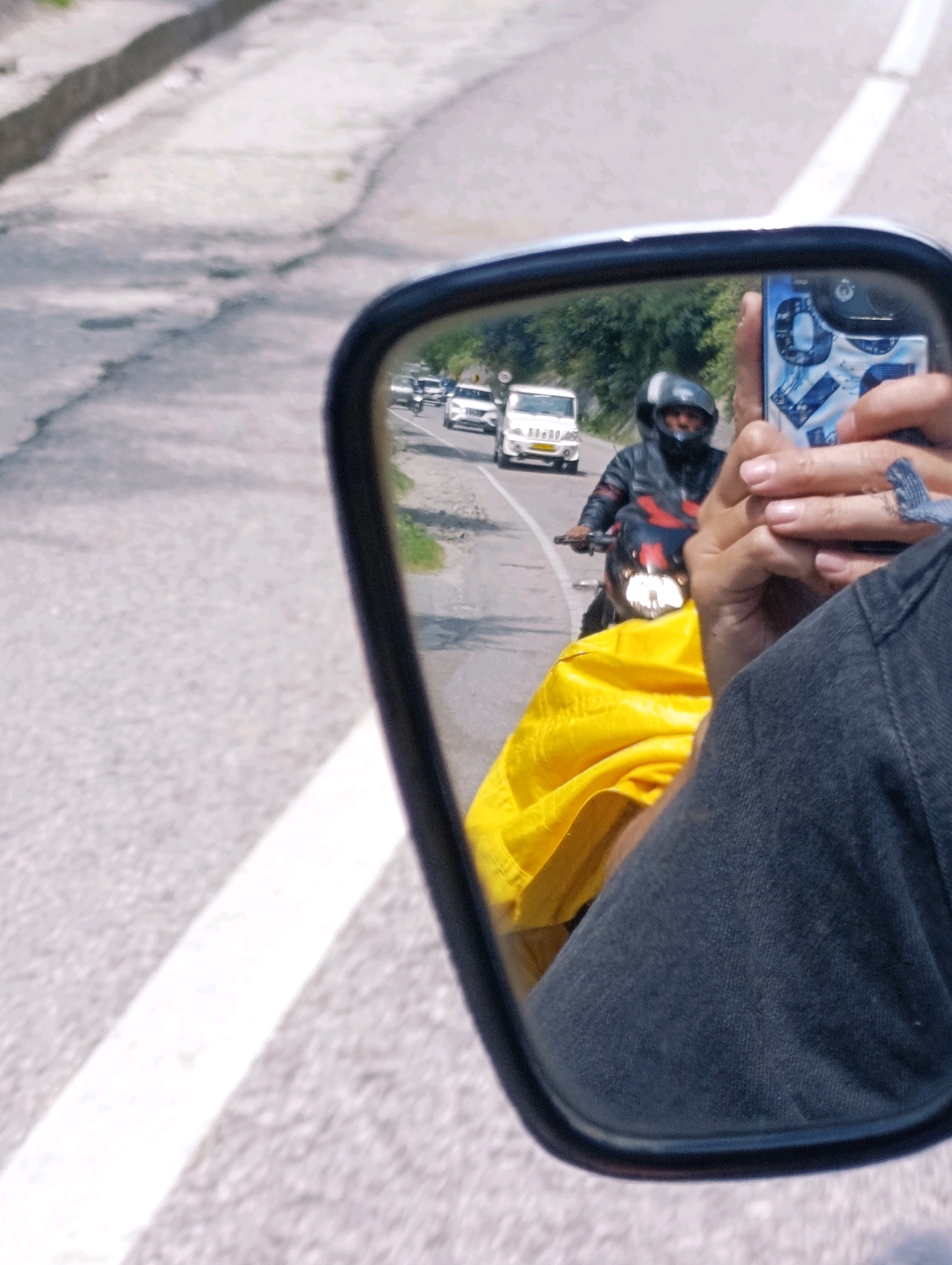 In the reflection of a motorcycle's side mirror, a man wearing a black helmet and a black and red jacket is riding a motorcycle. He is carrying a bright yellow tarp behind him. To his left and further ahead in the mirror's reflection, a white vehicle is visible. The scene appears to be on a paved road with a white line marking the lane. The background includes trees and a blurred landscape. The image's foreground shows the hand of a person holding a smartphone, which is capturing the reflection. This is a view from a motorcycle on a road in Chattan Seri, India.