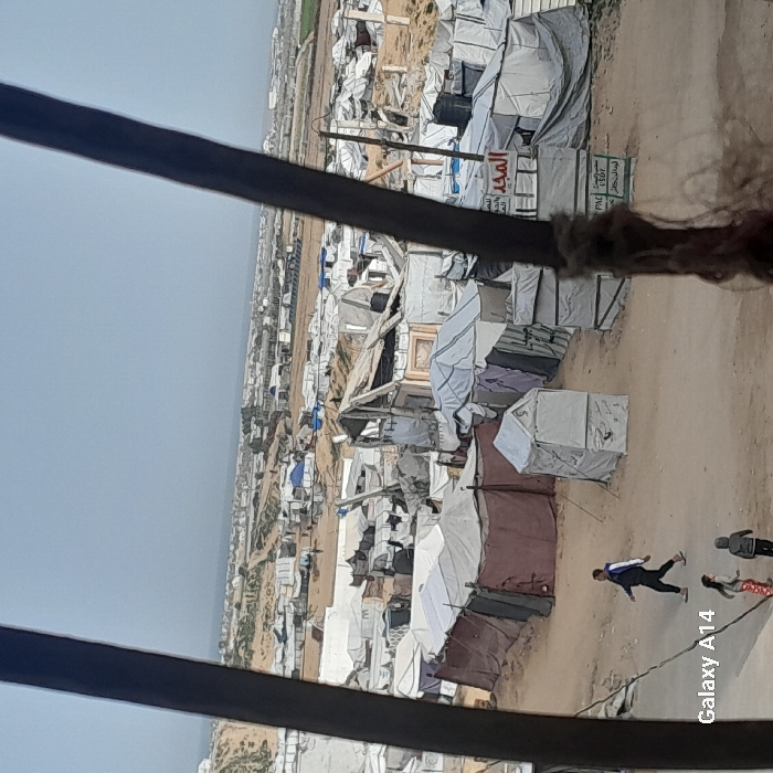 An expansive refugee camp is depicted, filled with densely packed tents of various colors and materials. The landscape is arid and dusty. In the foreground, figures can be seen walking along a dirt path winding through the camp. The photo is taken from an elevated perspective, looking down upon the camp. A blurred foreground object partially obscures the view.