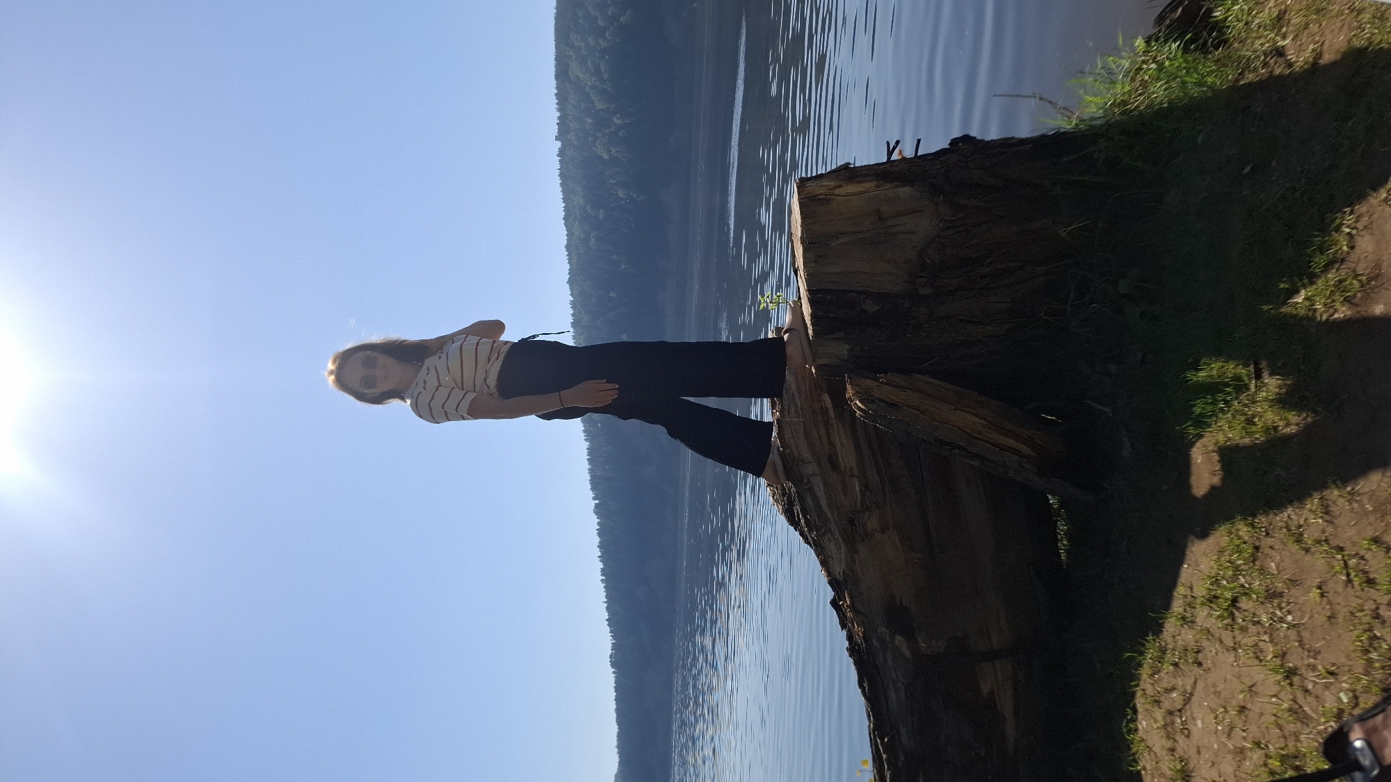 A young woman stands barefoot on a large, decaying tree stump near a body of water, likely a lake, under a bright, clear sky. The location appears to be rural or natural, with a wooded area across the lake. The woman is wearing dark pants and a light-colored striped shirt. The time of day appears to be midday, given the position of the sun. The location is near Noviy, Russia based on the provided coordinates.
