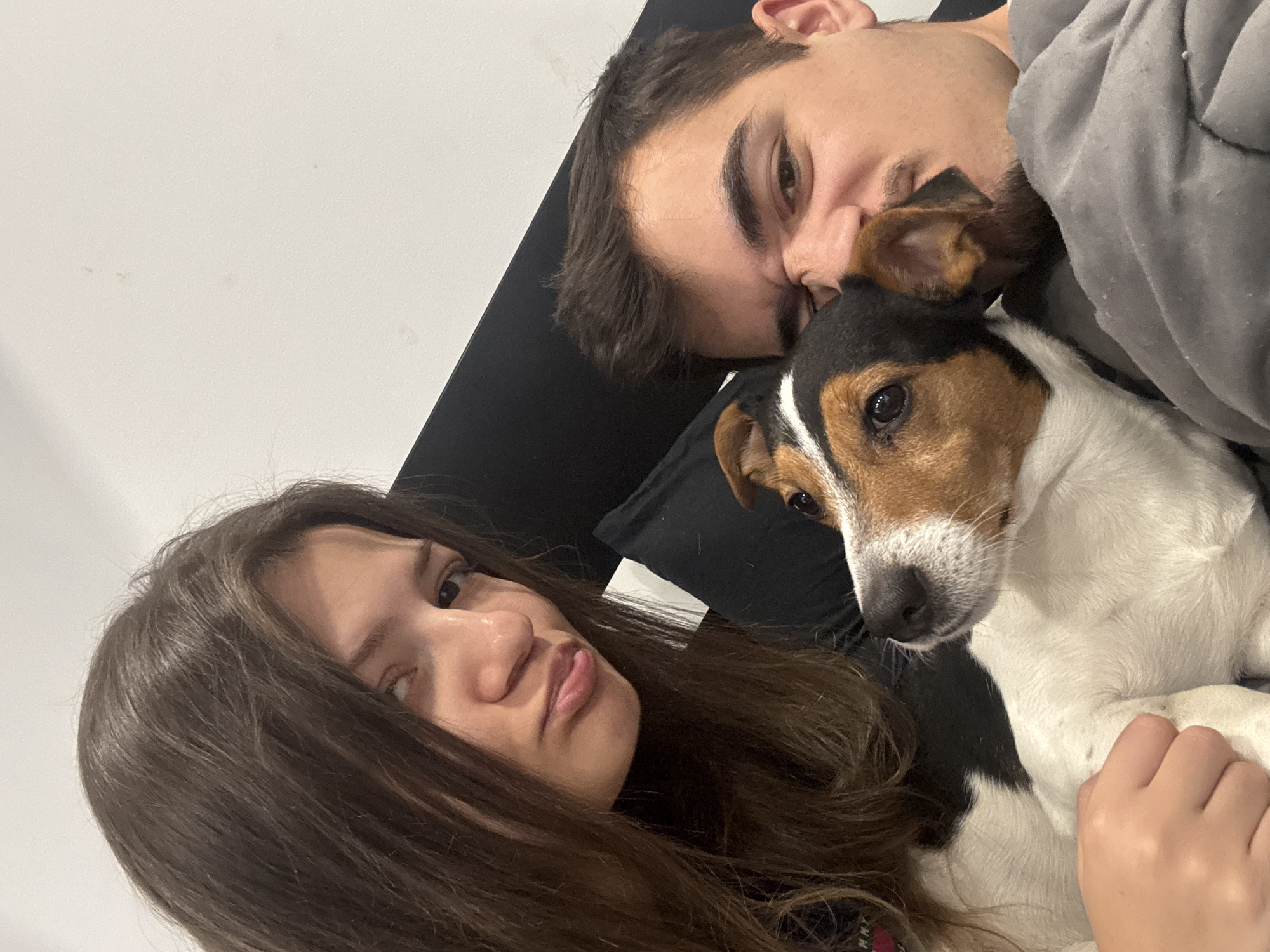 The image shows a selfie of a young woman, a young man, and a dog. The woman has long, dark hair and is looking at the camera with a neutral expression. The man is lying next to her with his head resting near the dog's head. The dog is a Jack Russell Terrier with brown and white fur. They are all lying on a dark-colored surface, possibly a bed or couch. The background is a plain white wall. It is not possible to ascertain the location in Sofia, Bulgaria, from the image alone.