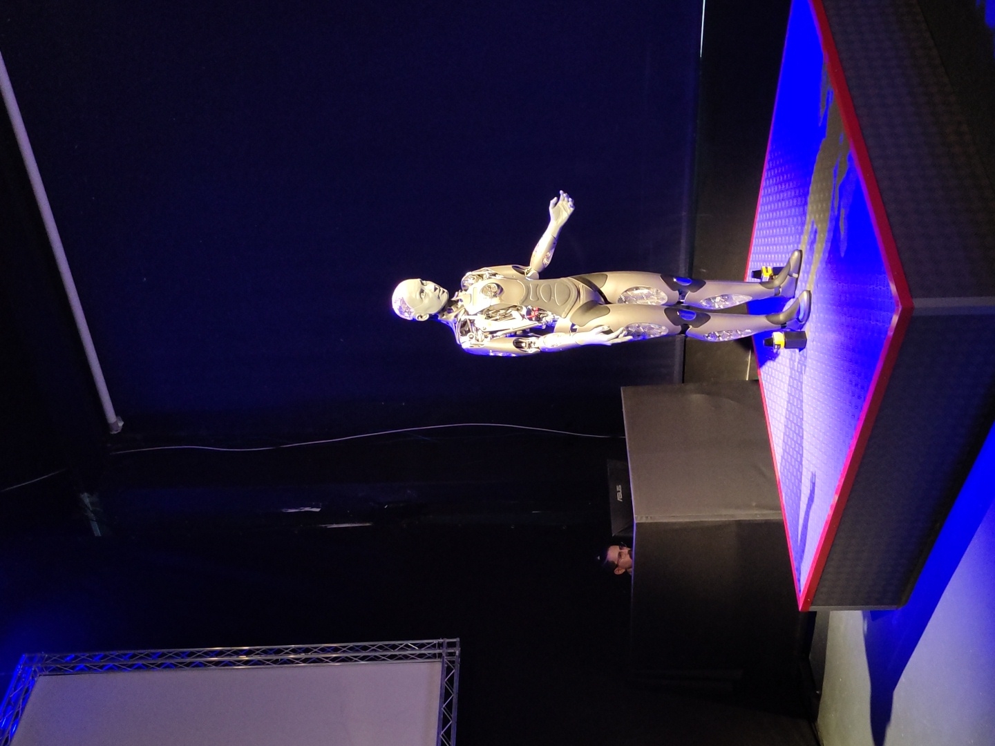An advanced humanoid robot stands on a brightly lit, blue-toned platform. The robot is silver with visible internal workings and articulated joints, and it is positioned in the center of the frame with its right arm extended slightly to the side, palm up, in a gesture that could be interpreted as an invitation or a presentation. To the left of the robot, partially obscured by a black structure, a person is visible behind a computer monitor. The background consists of a dark stage with a large projection screen on the left and a dark wall or backdrop on the right, illuminated by a blue stage light. The scene appears to be an indoor event or presentation, likely at an exhibition or a technology demonstration, potentially in Haifa, Israel.