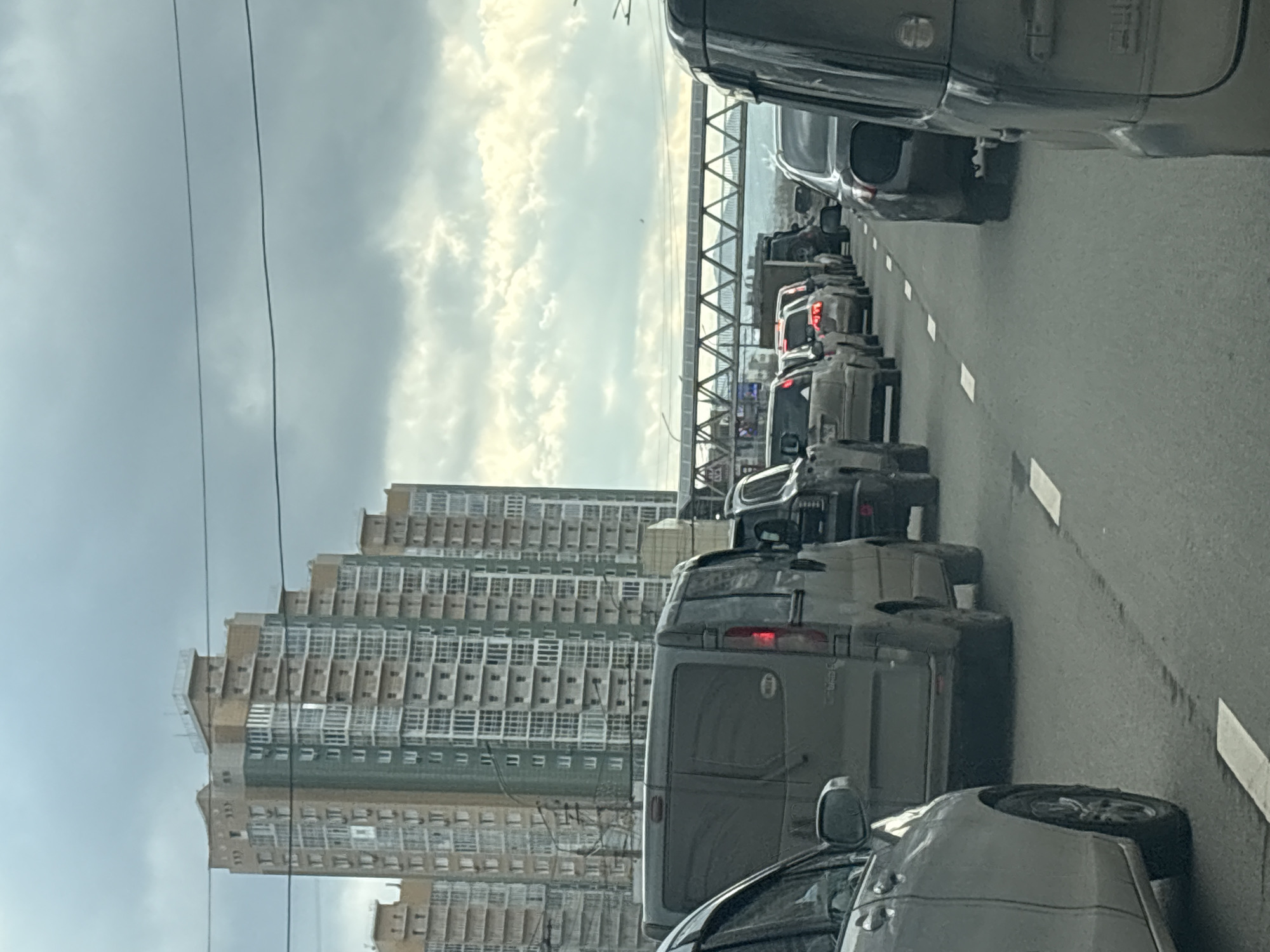 The image depicts a daytime traffic jam on a multi-lane road in an urban environment, likely a bridge or overpass. Numerous cars, including sedans, SUVs, and a van, are seen bumper-to-bumper, suggesting slow or stopped movement.
In the background, a large, modern residential building with many windows and balconies stands prominently to the left. Further in the distance, behind the traffic, a distinctive bridge-like structure with a lattice design is visible, beyond which appears to be a wide body of water under a bright, cloudy sky. Overhead, a few power lines cut across the frame.
The weather is bright with significant cloud cover, suggesting either morning or late afternoon light. While no individuals are directly visible, the presence of many vehicles implies a busy urban setting. The overall scene conveys a sense of urban congestion and waiting. There is no discernible text visible within the image. Given the provided context, the scene is located in Krasnoyarsk, Russia.
