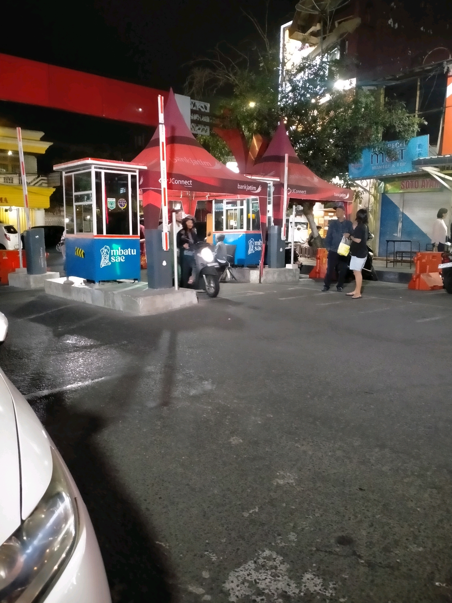 This is a nighttime, medium-shot photograph of a toll booth area, likely in the city of Batu, Indonesia, given the presence of the "mbatu sae" branding. The scene is set outdoors on a paved surface, which appears wet and reflective, suggesting recent rain.
The central focus is a modern toll booth with a glass-enclosed operator's booth and red and white barrier arms. A blue box with yellow chevron markings and the "mbatu sae" logo is prominently displayed on the left side of the booth. To the right of the booth, a red tent with "bankjatim" and "JConnect" branding is set up.
Several individuals are visible. A person on a scooter is stopped at the toll booth, with their face obscured by a helmet and the lighting. To the right of the booth, a man in a dark jacket and cap stands talking to a woman wearing a dark top and light-colored shorts, holding a yellow bag. Further to the right, another person in a light-colored jacket is visible.
In the background, there are buildings with illuminated signs, including a red overhead structure and a blue sign with "M&T" and "MANTAP & TOP" text. The overall environment suggests a commercial or service area, possibly a parking lot or a main thoroughfare with toll collection. The lighting is artificial, casting shadows and creating reflections on the wet ground.